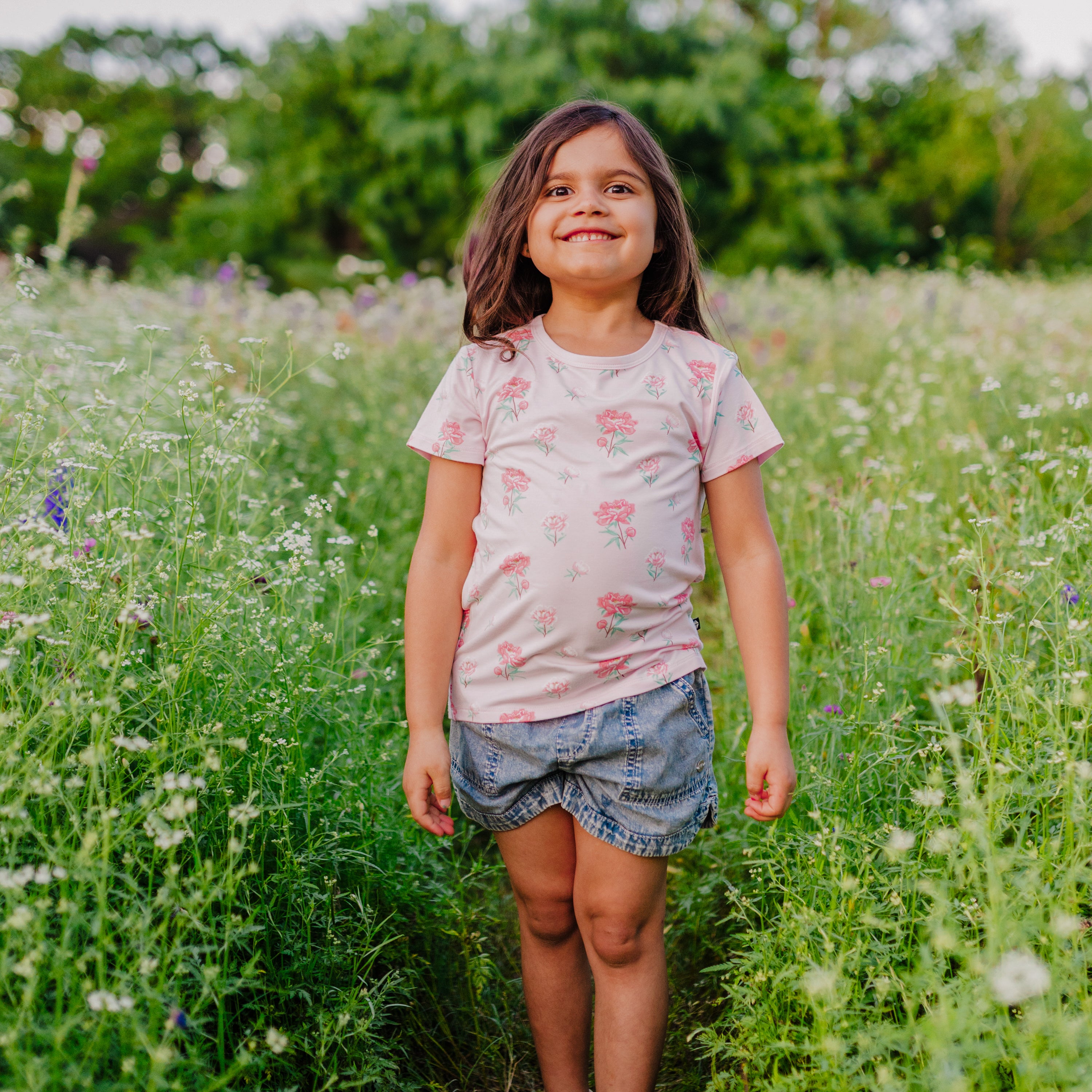 Smiling girl model wearing the Toddler Basic Tee in Sakura Peony with jean shorts standing in a field of wild flowers