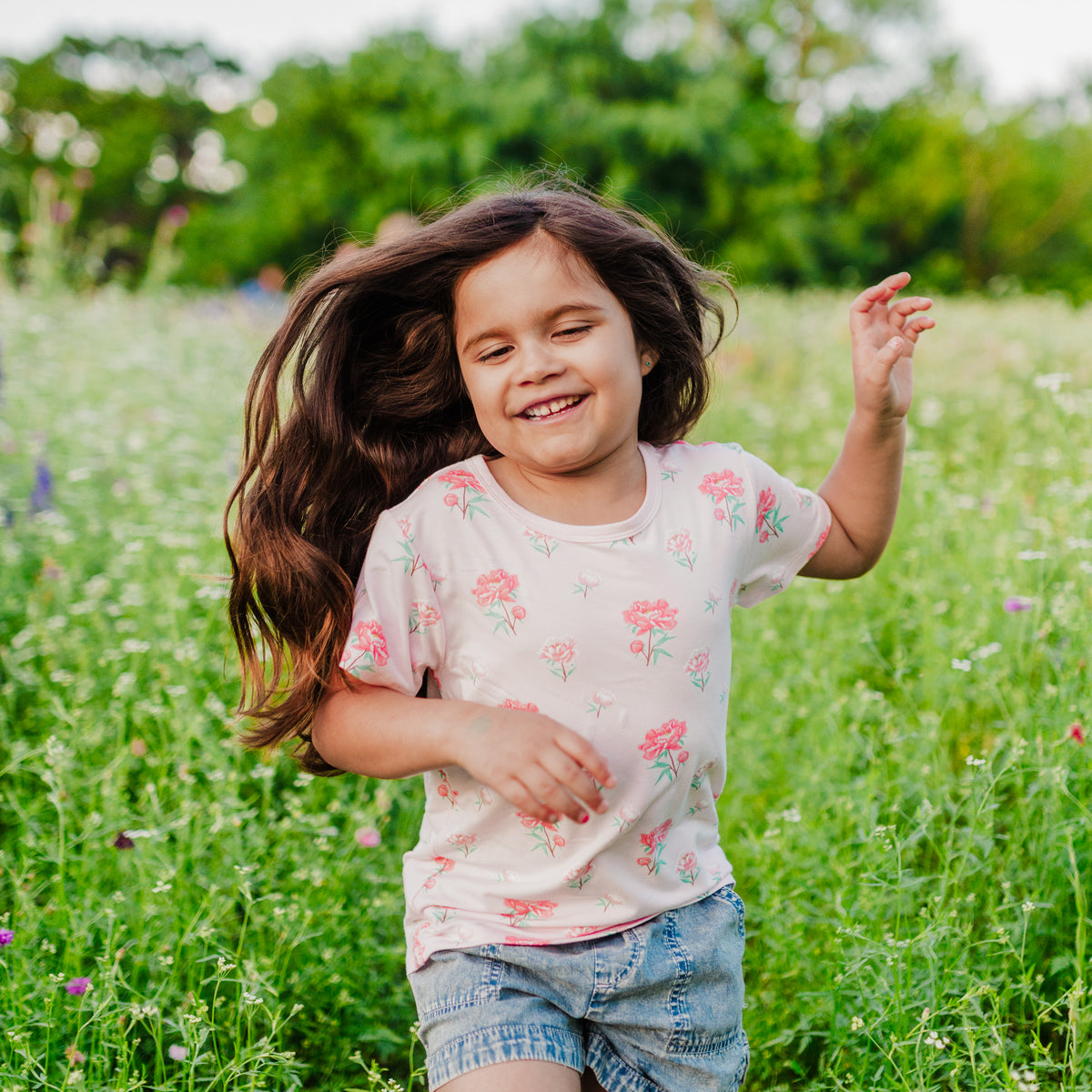 Close up of young girl smiling wearing the Toddler Basic Tee in Sakura Peony paired with jean shorts walking through a wild flower field