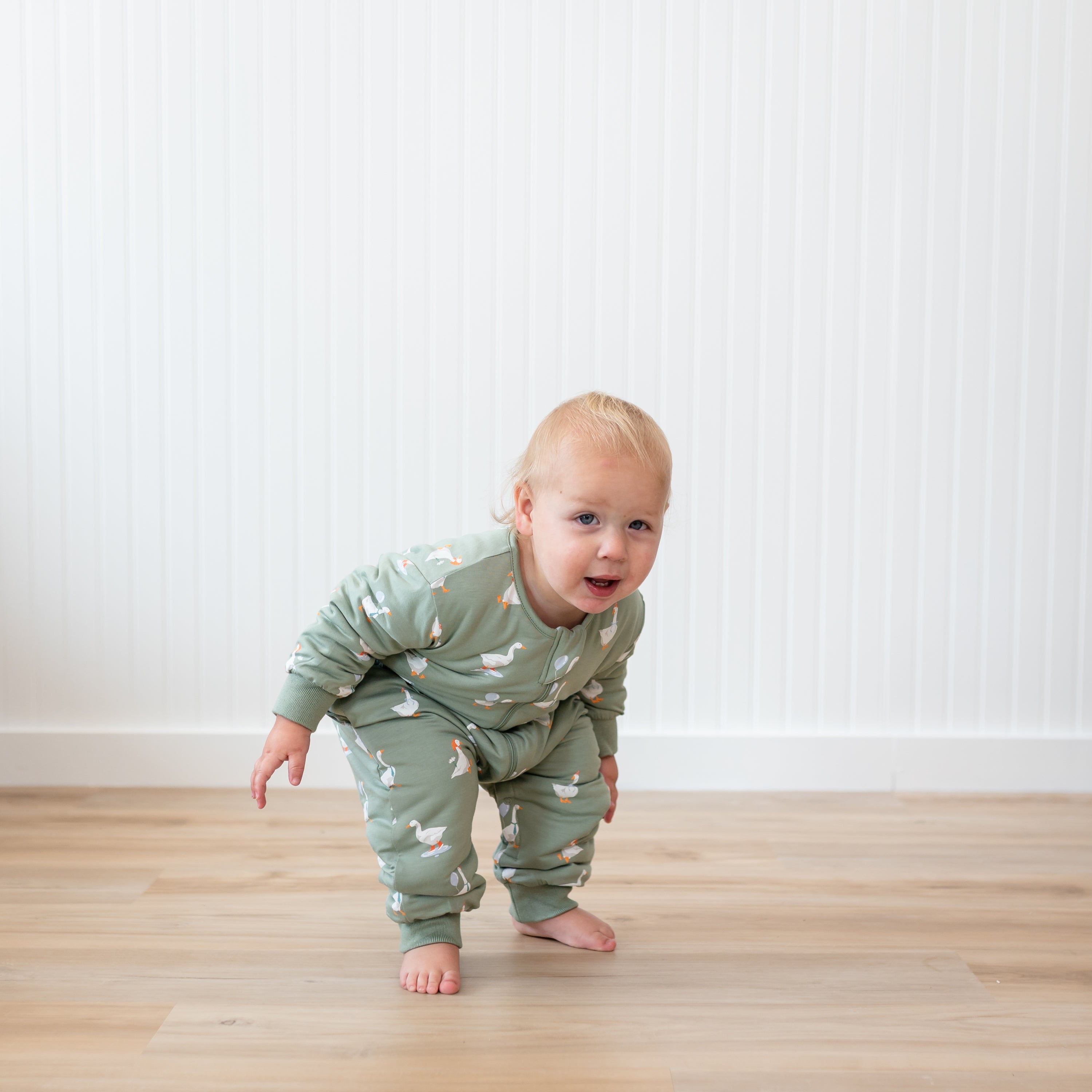 Toddler crouching wearing the Cozy Playsuit in Silly Goose in front of a white paneled wall