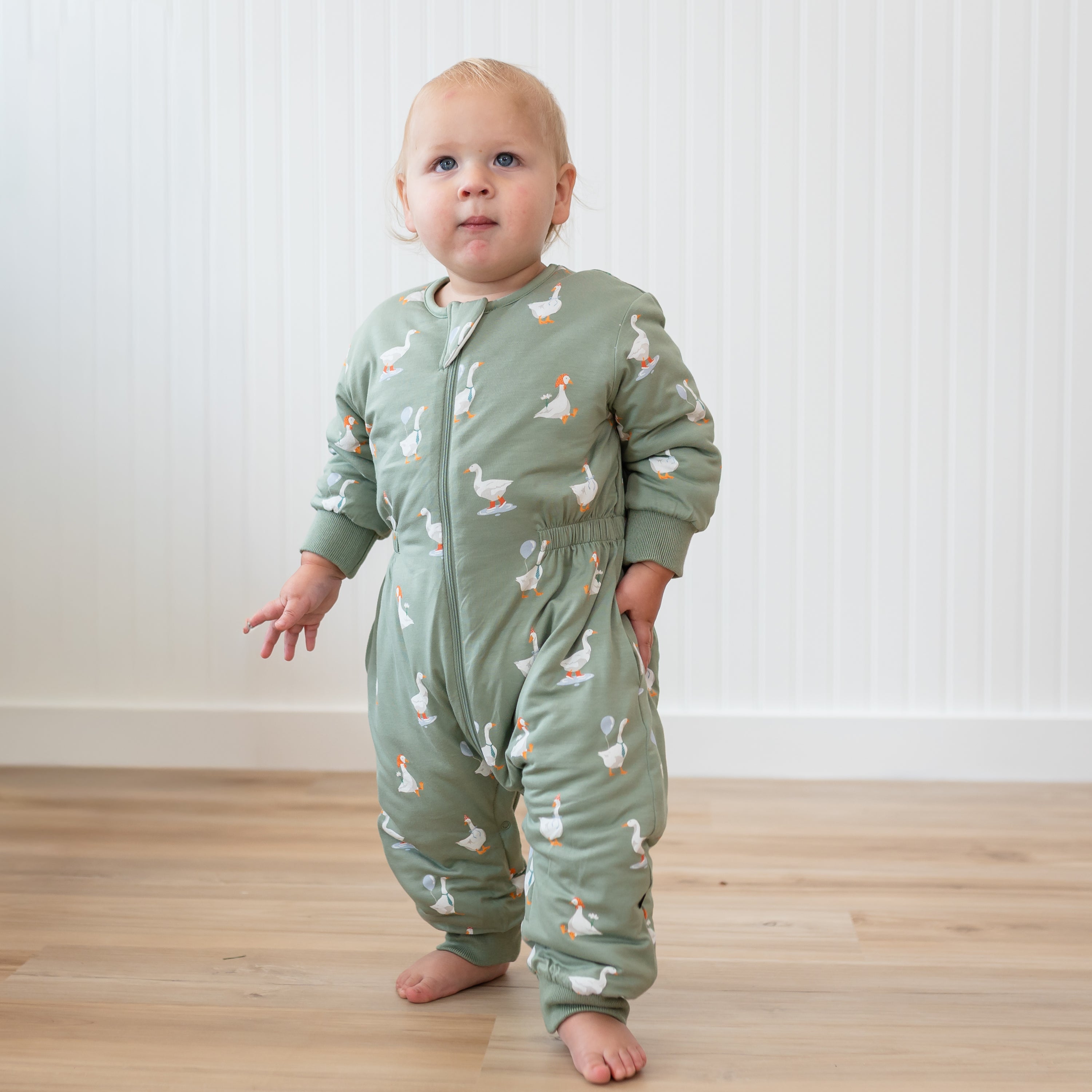 Toddler standing wearing the Cozy Playsuit in Silly Goose standing in front of a white paneled wall