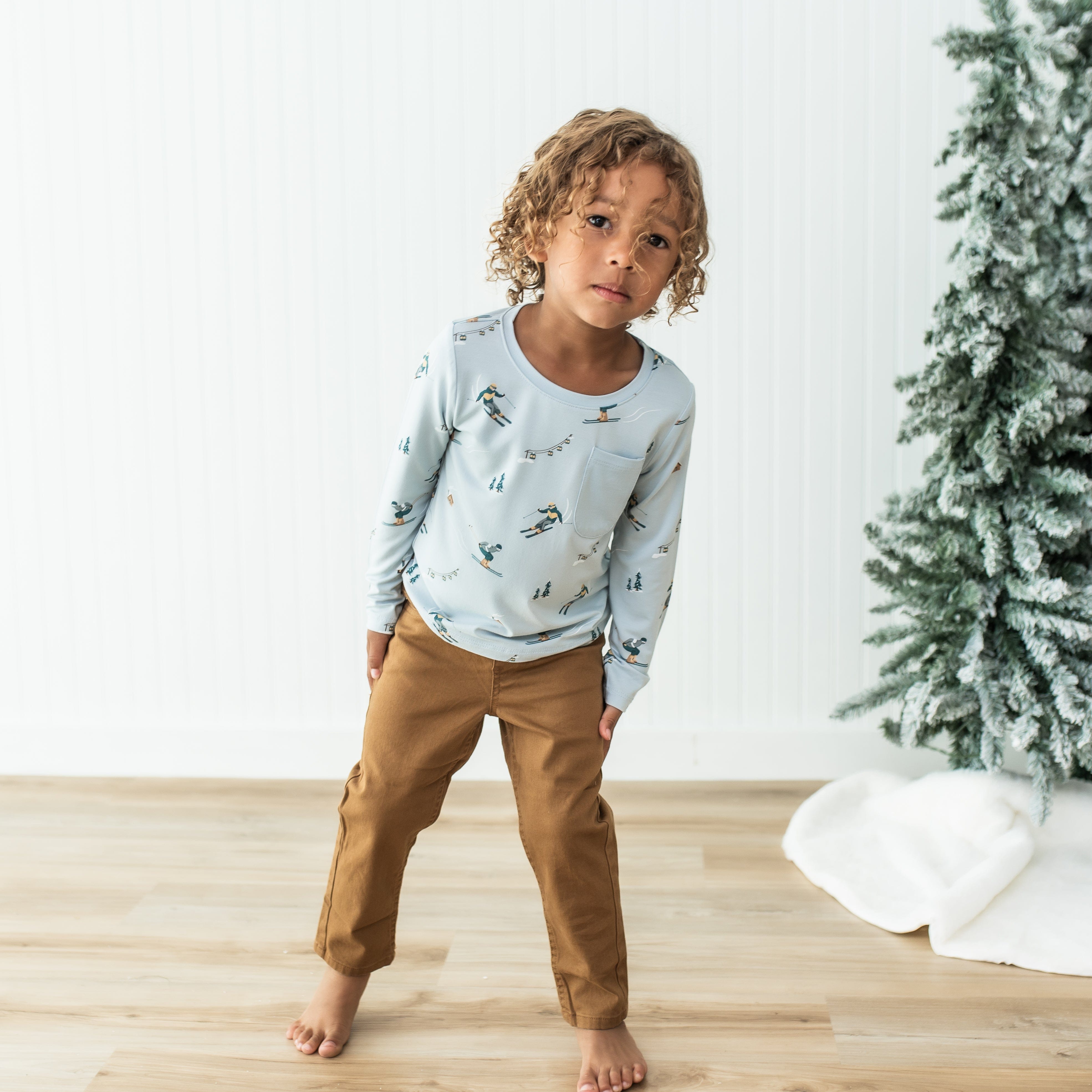 Young boy standing in front of a white paneled wall wearing the Long Sleeve Toddler Crew Neck Tee in Ski paired with bronze colored pants