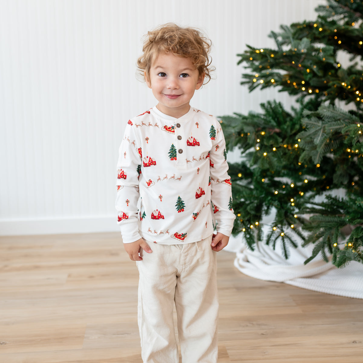 Young boy standing beside a Christmas tree wearing the Long Sleeve Toddler Henley Tee in Santa Sleigh and light colored pants
