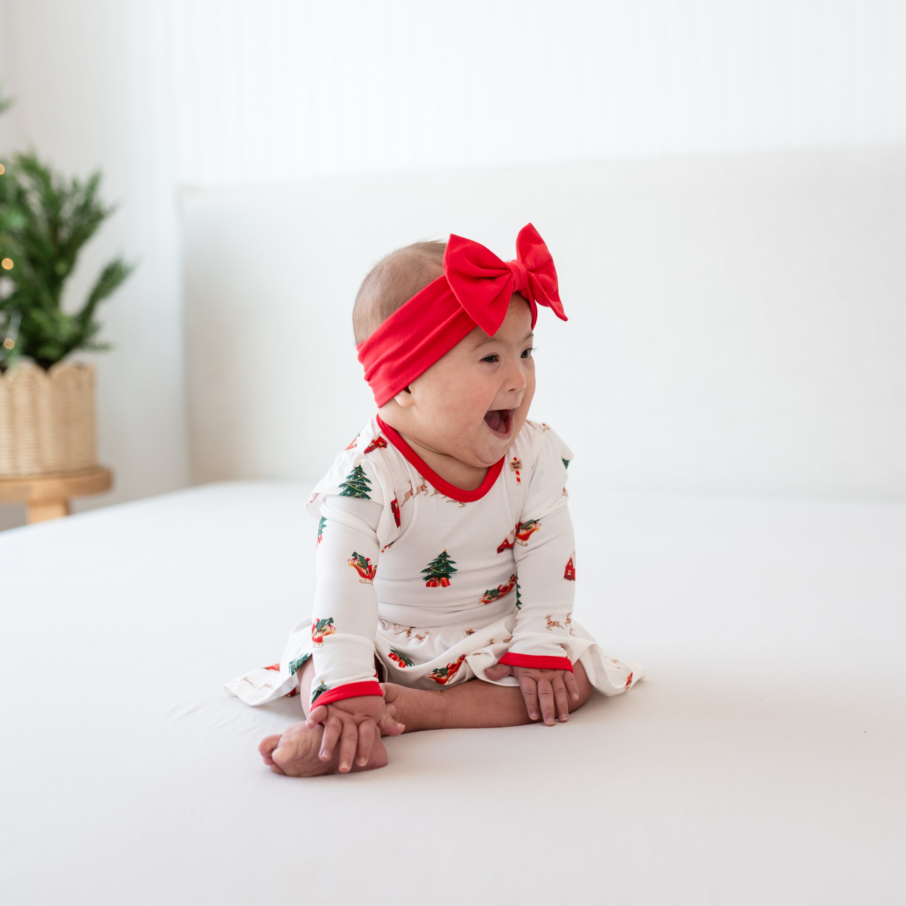 Young infant sitting on a bed wearing the Long Sleeve Twirl Bodysuit Dress in Santa Sleigh and Cardinal bow headband