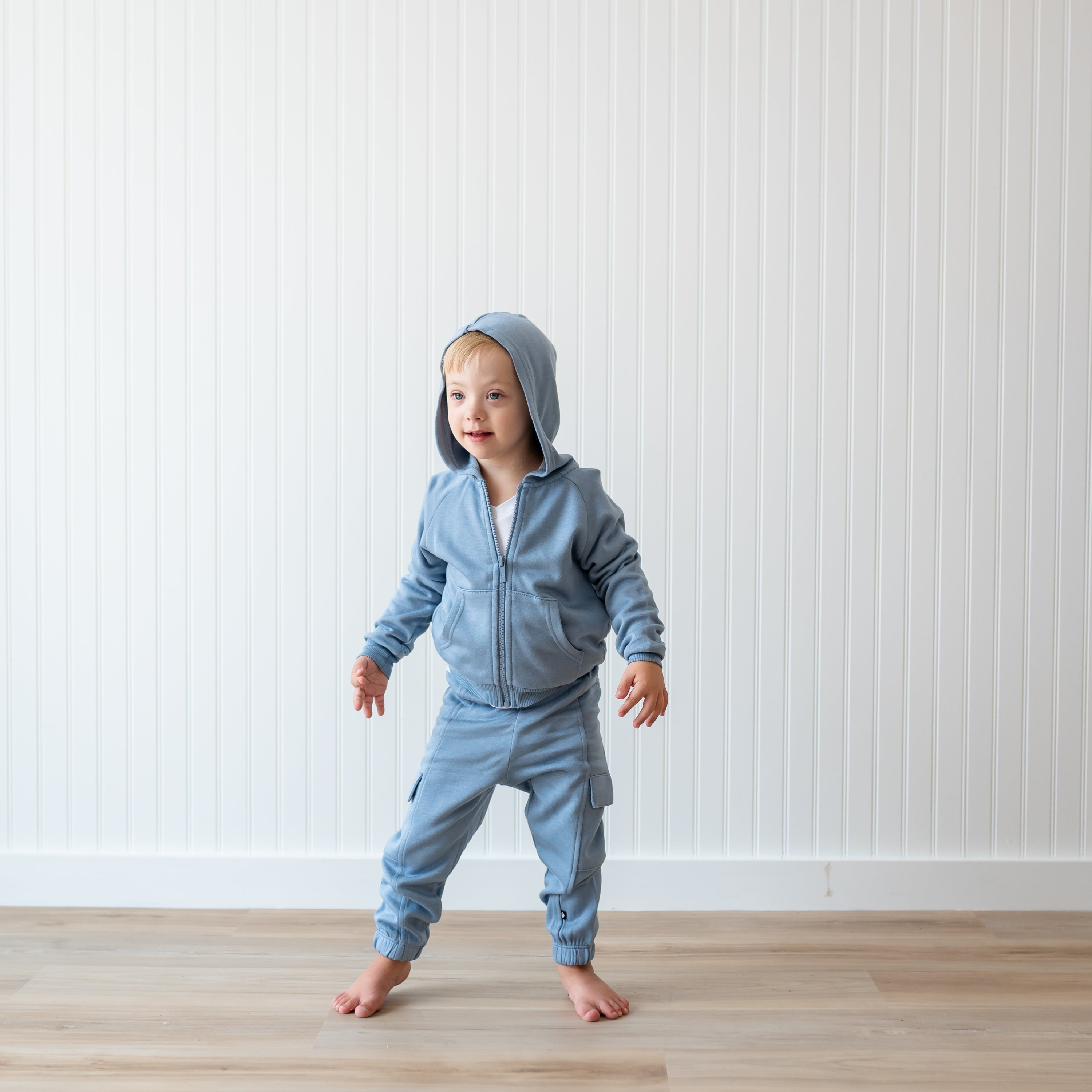 Young boy standing in front of a white paneled wall wearing the Fleece Zip Up Hoodie in Slate and matching fleece cargo pant