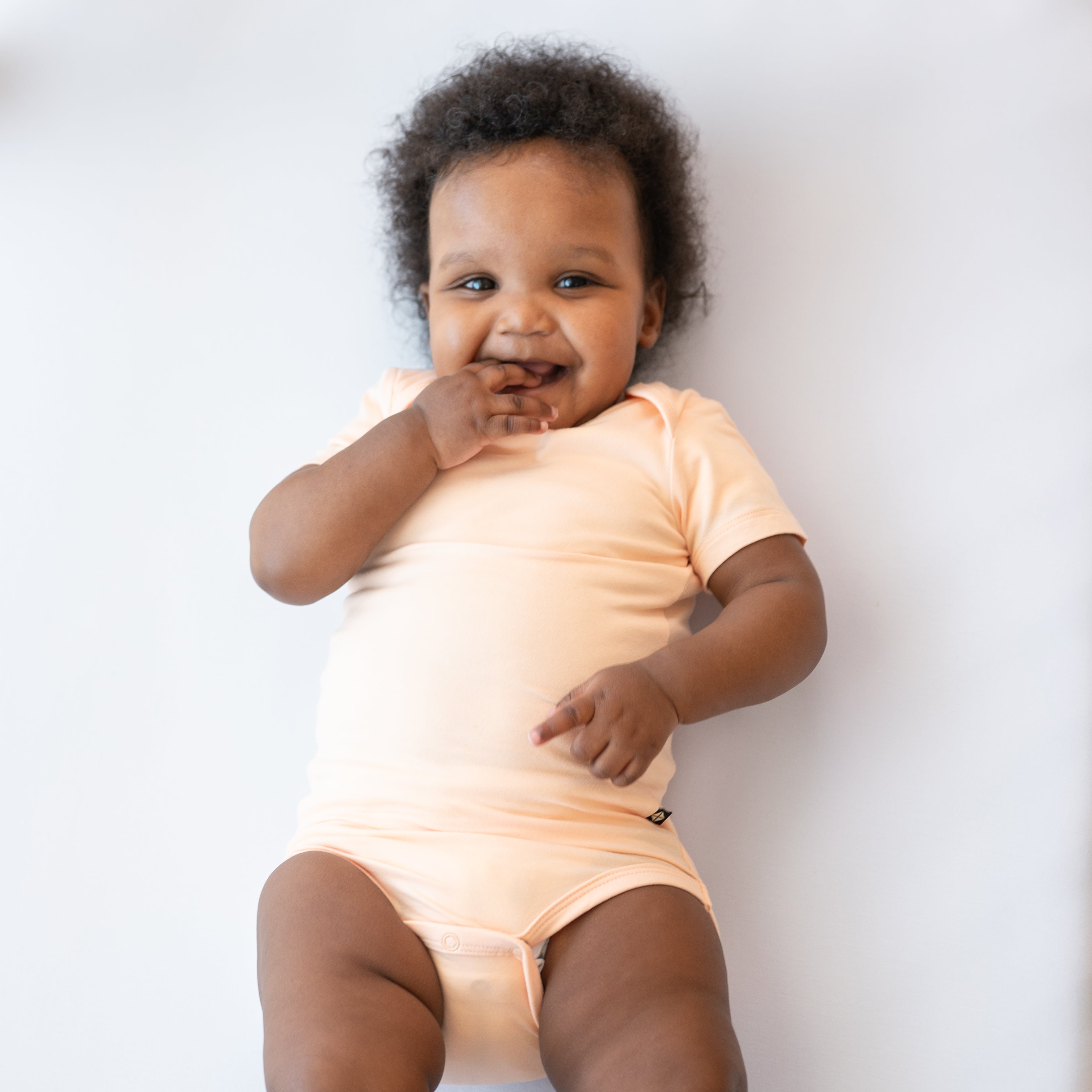 Smiling baby laying in a crib with hand in their mouth wearing the Bodysuit in Canyon