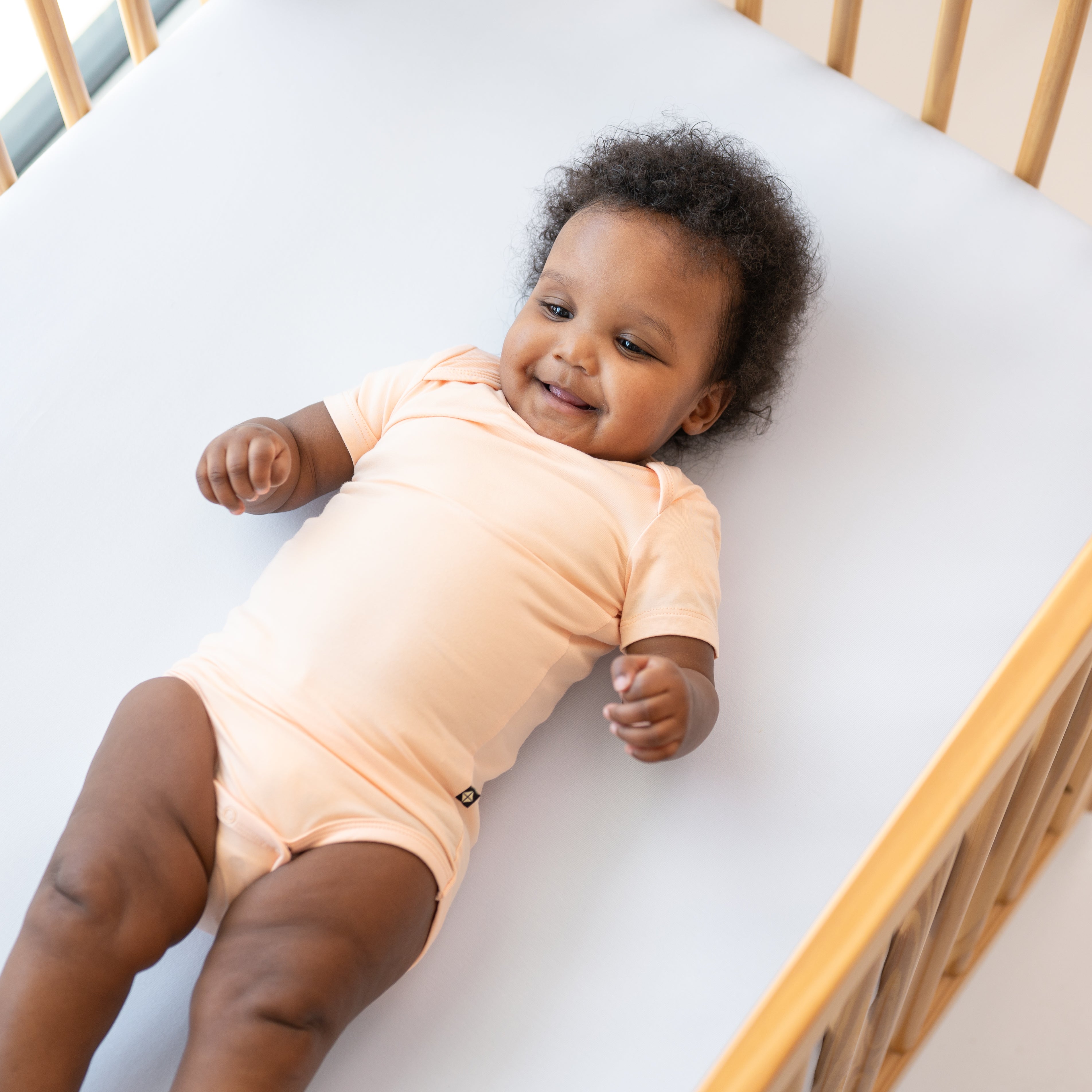 Smiling baby laying in a crib wearing the Bodysuit in Canyon