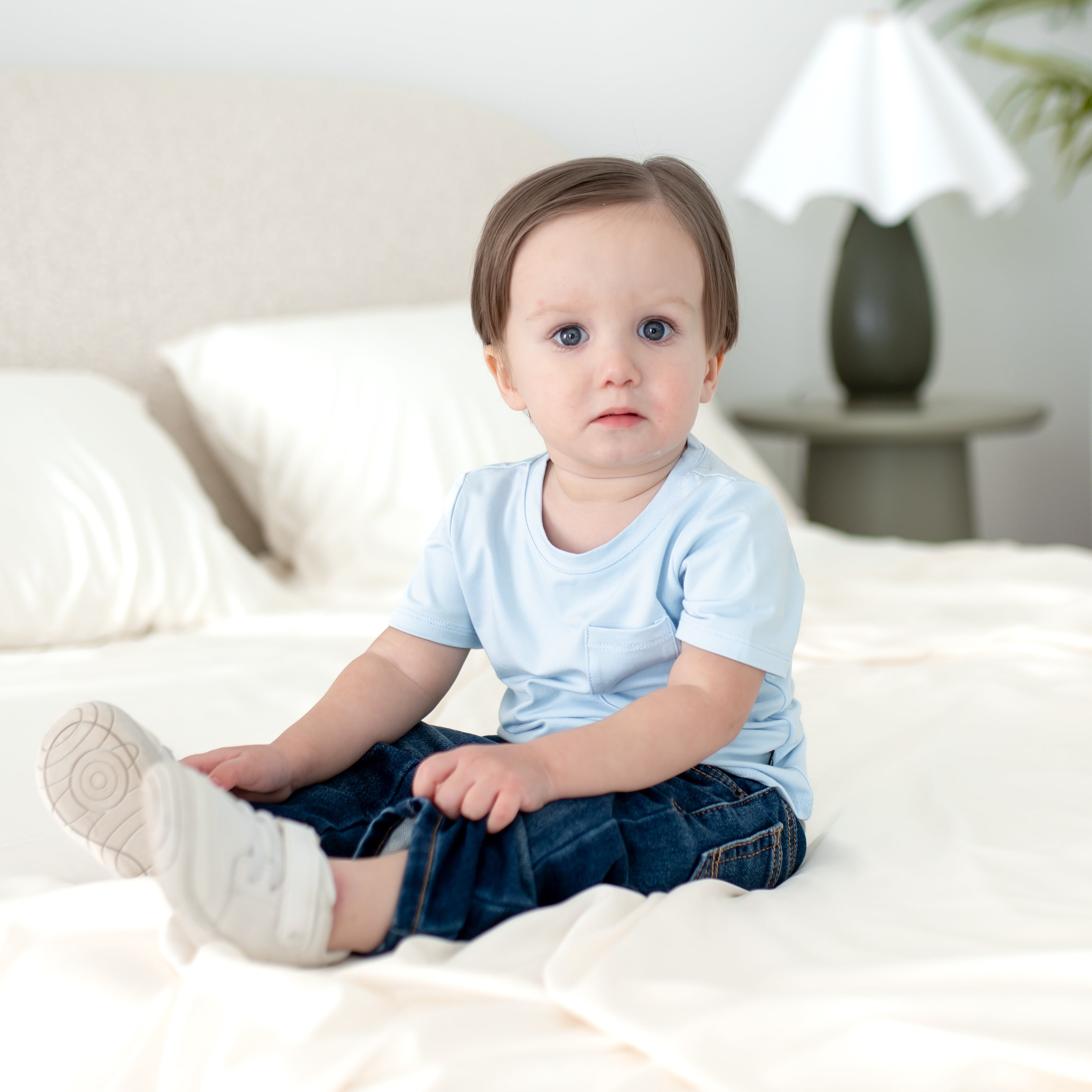 Young toddler sitting on a white bed wearing the Toddler Crew Neck Tee in Breeze with dark wash jeans