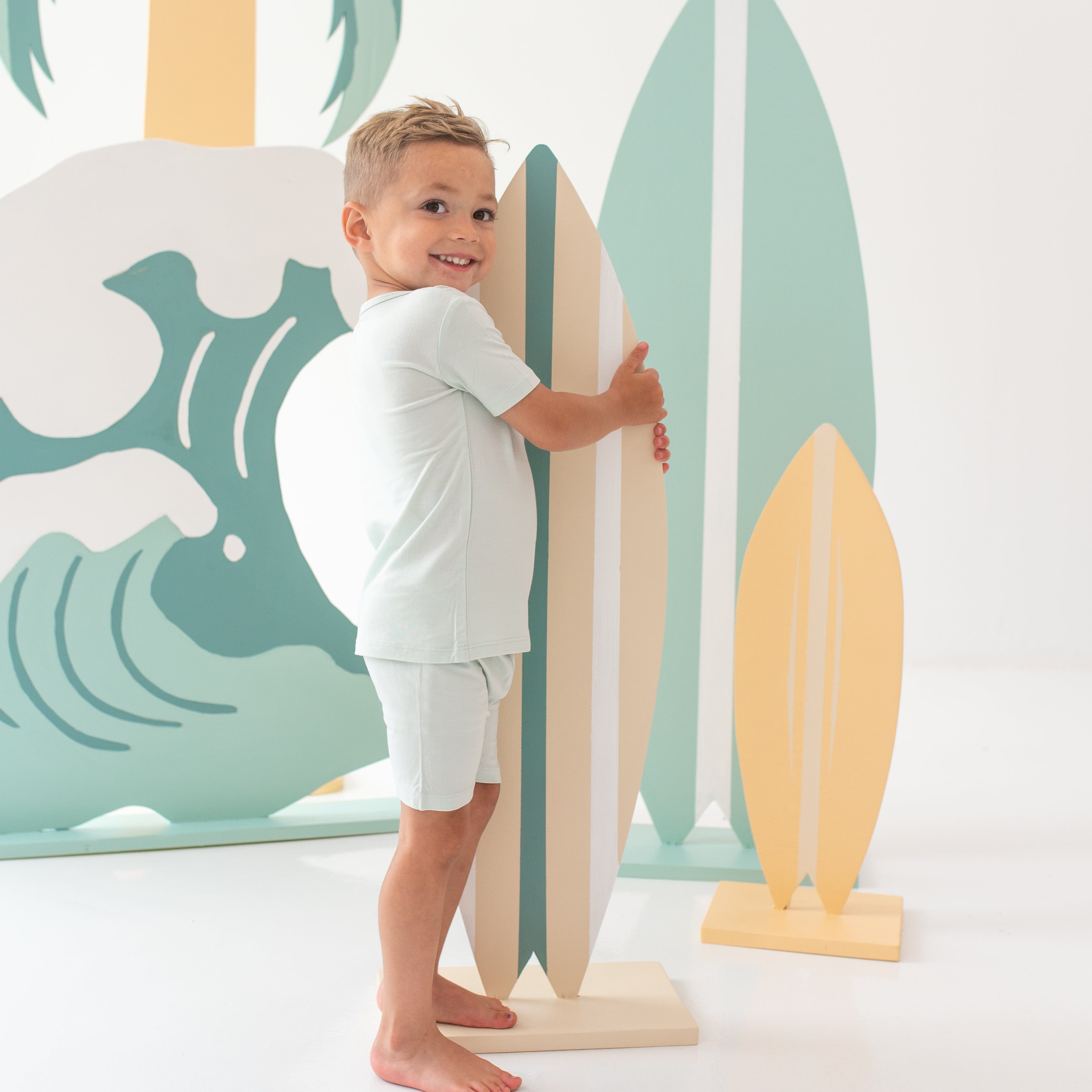Young boy wearing the Short Sleeve Pajamas in Dew holding a wooden surf board prop