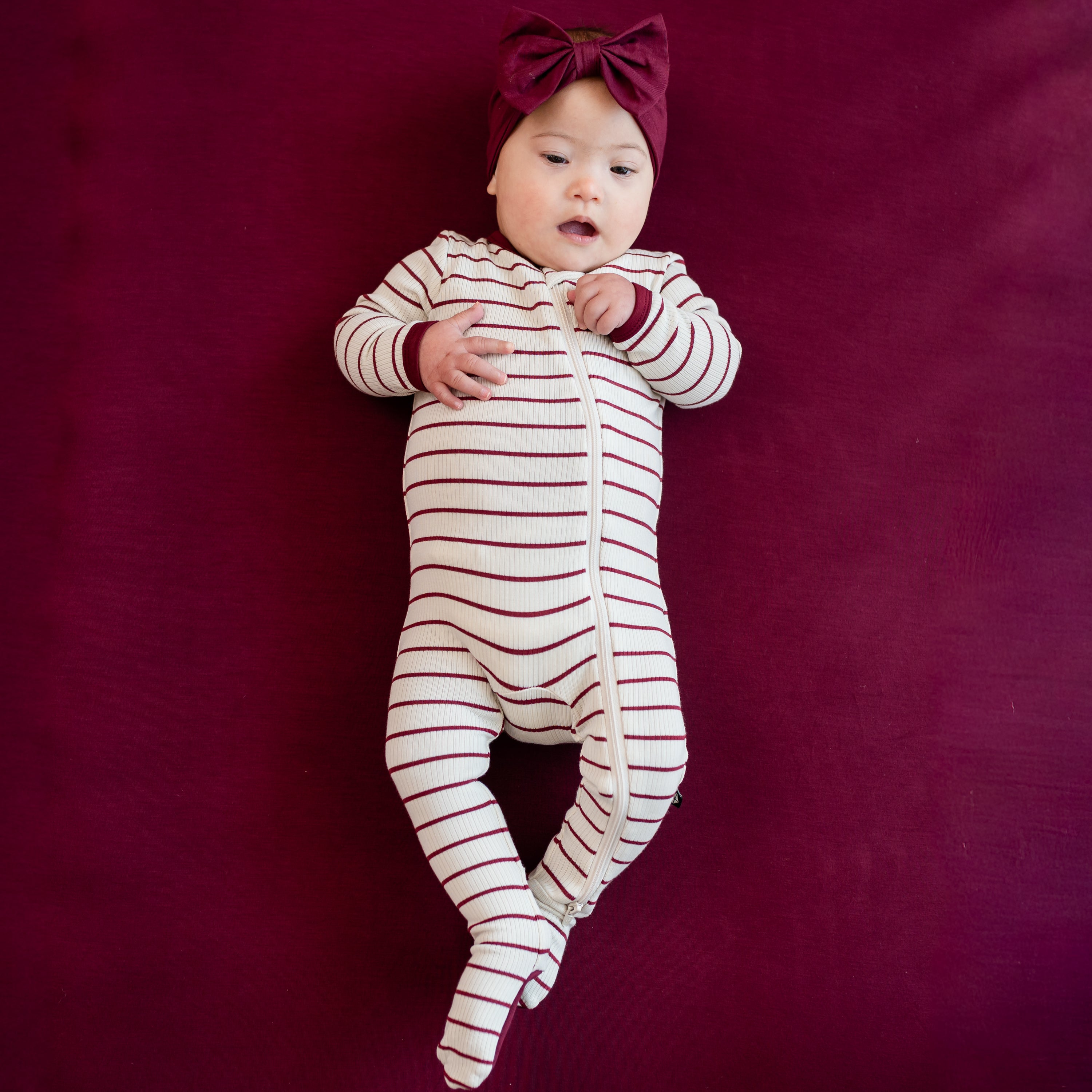 Infant laying on a Burgundy crib sheet wearing the Ribbed Zipper Footie in Burgundy Stripe with burgundy bow headband