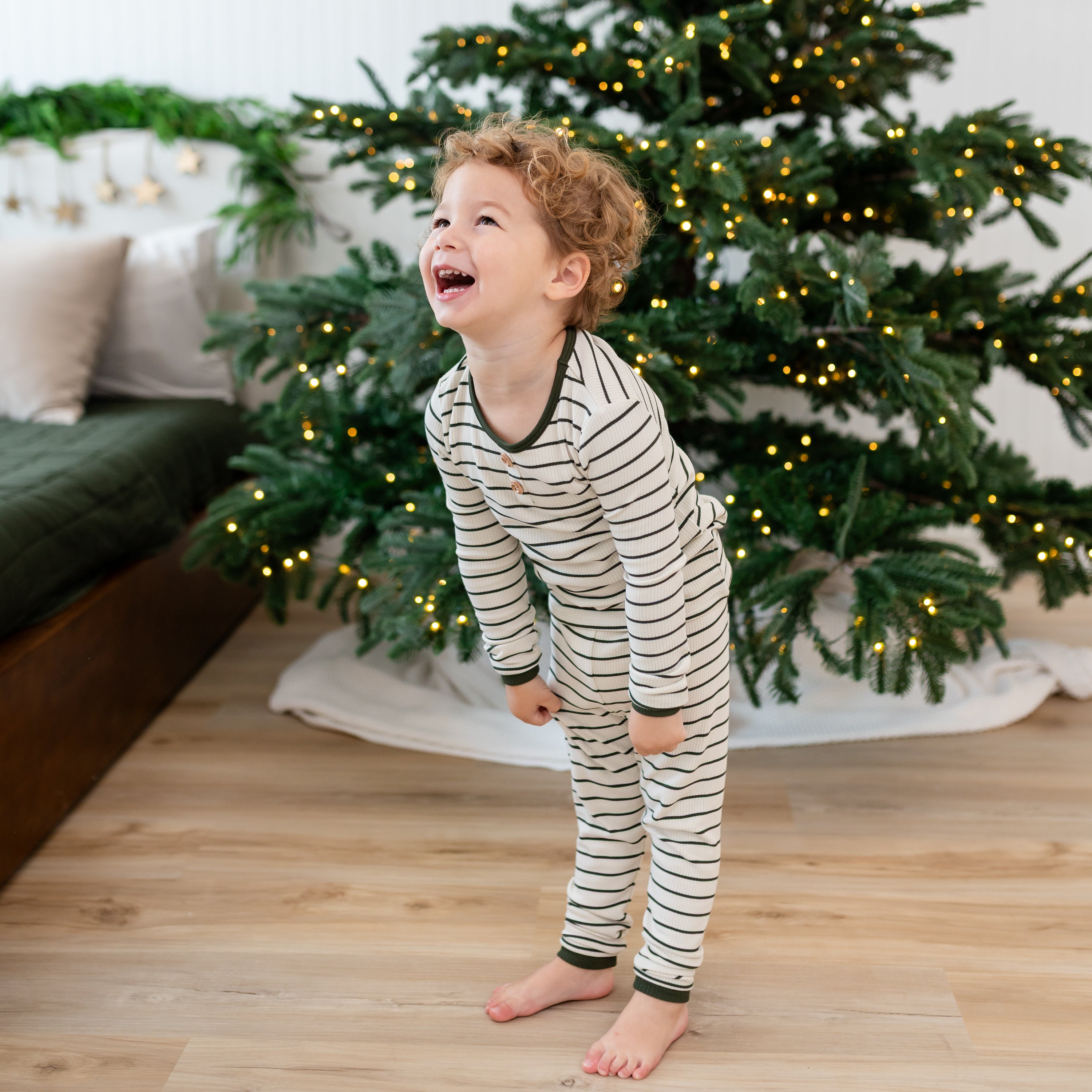 Smiling toddler standing in front of a lit Christmas tree wearing the Ribbed Henley Set in Fir Stripe