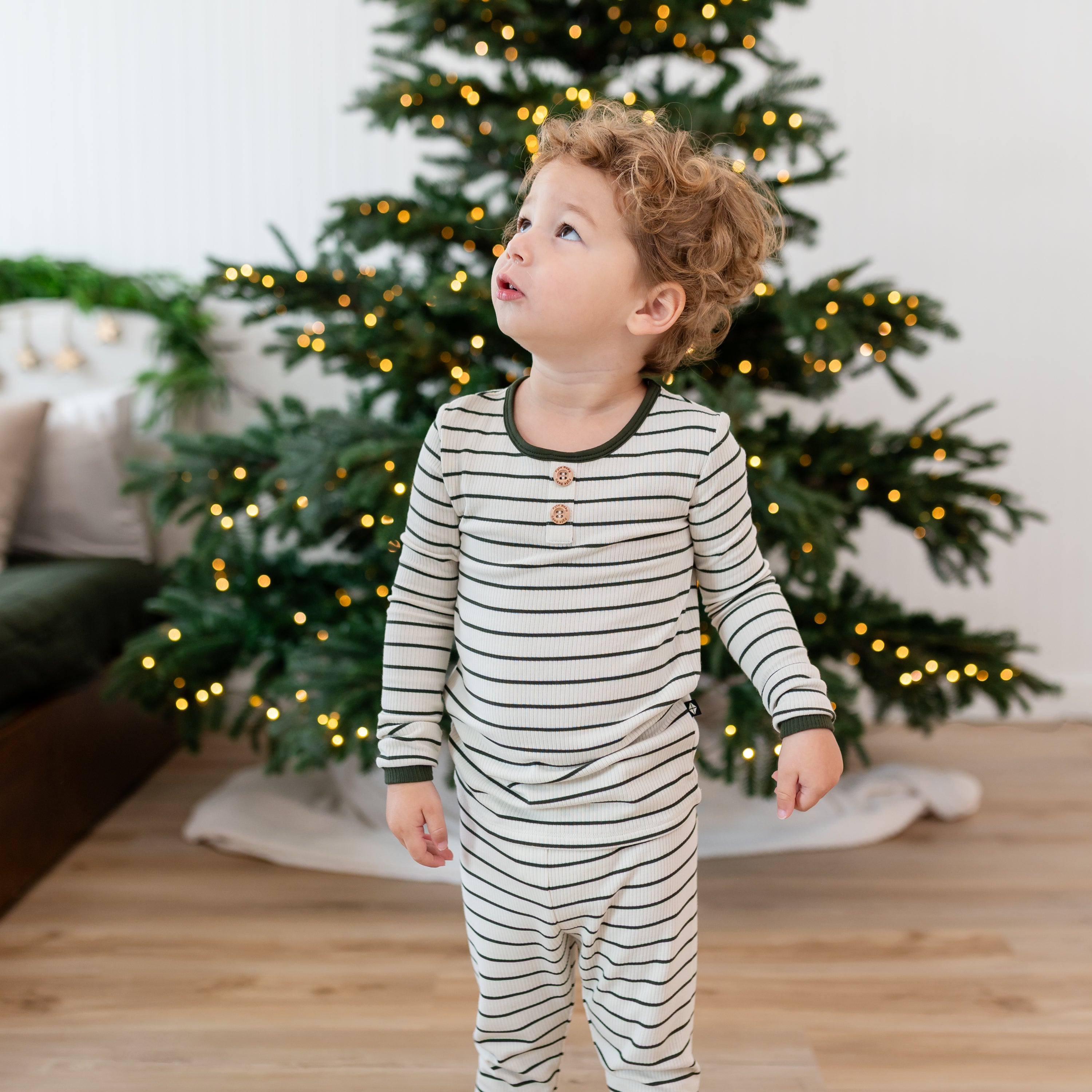 Close up of the Ribbed Henley Set in Fir Stripe shown on a toddler looking up standing in front of a lit Christmas tree
