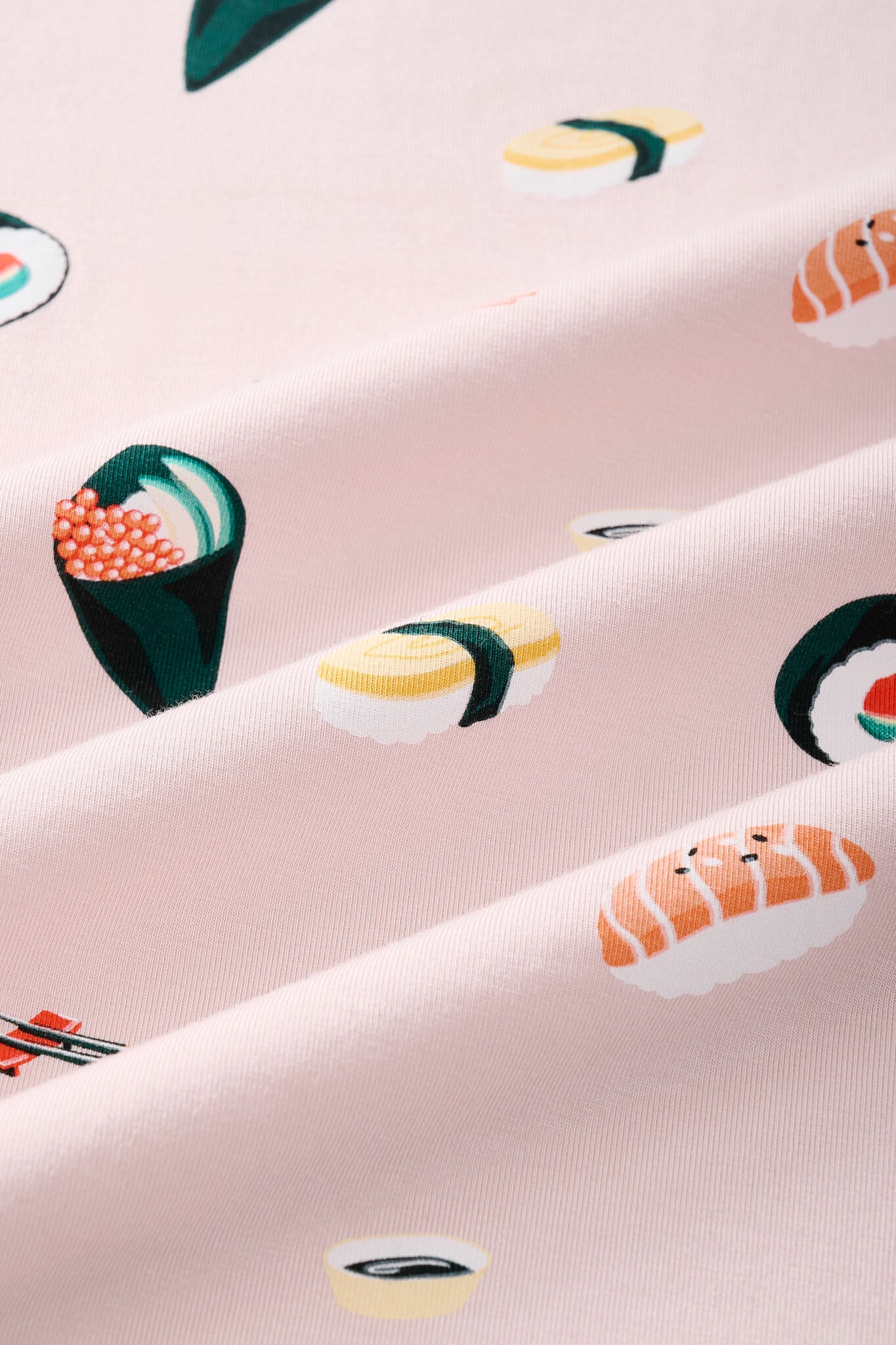 A close-up of a pink fabric with a repeating sushi print, including nigiri, maki, and sashimi motifs in yellow, green, orange, and white.