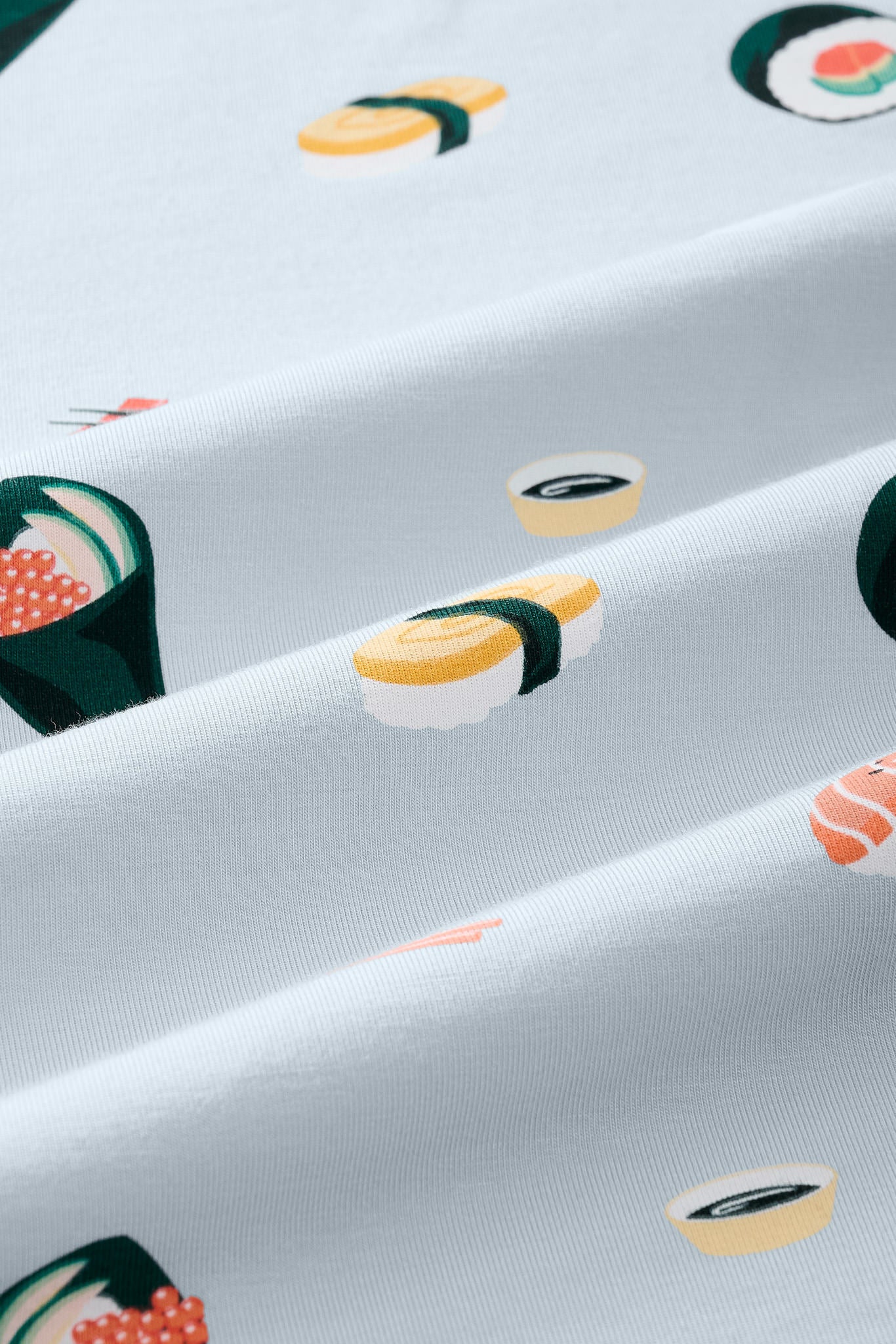 A close-up of a light blue-gray fabric with a repeating sushi print, including nigiri, maki, and sashimi motifs in yellow, green, orange, and white.
