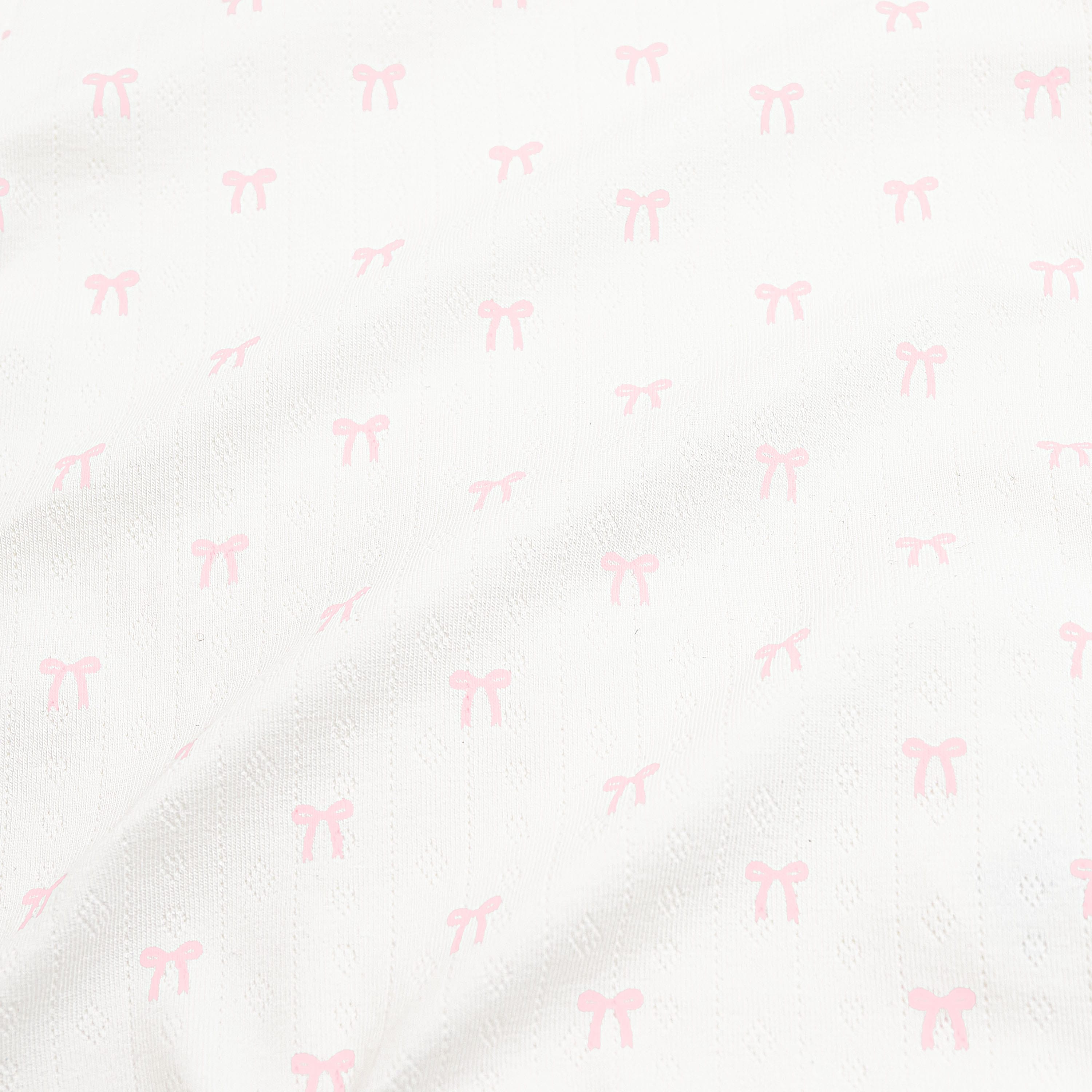 Close up of the pointelle fabric: An all-over print of dainty light pink bows over a white background.