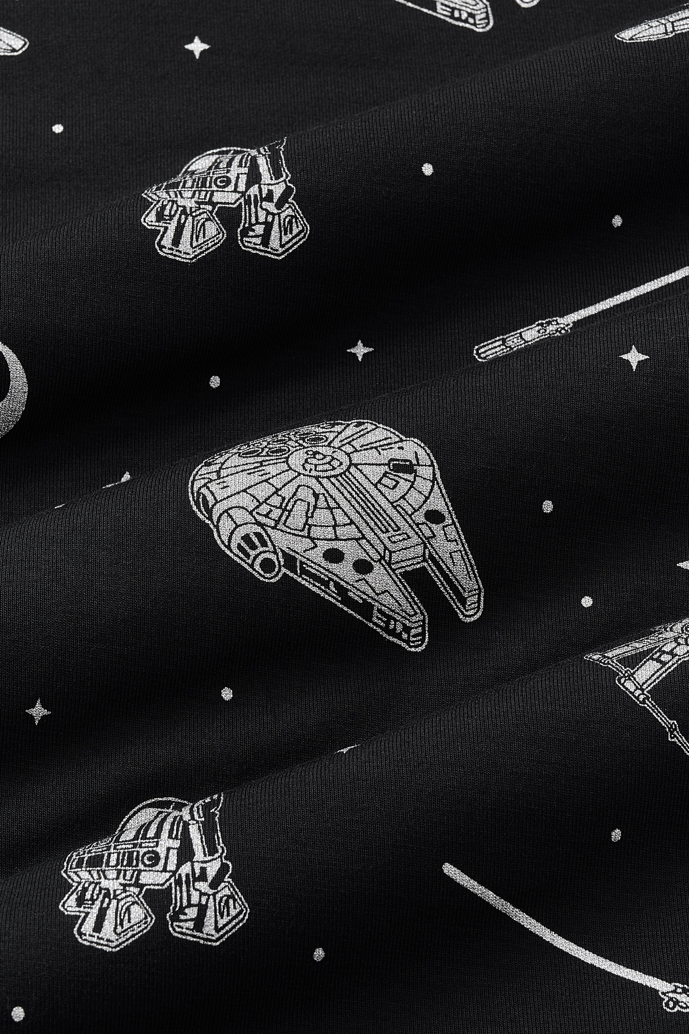 Close up of bamboo fabric: A repeating, monochromatic print featuring: a grey Milennium Falcon; a grey X-wing starfighter; a grey R2-D2; a grey lightsaber; a grey Rebel Alliance starbird symbol; and various grey twinkles and dots, over a black background.