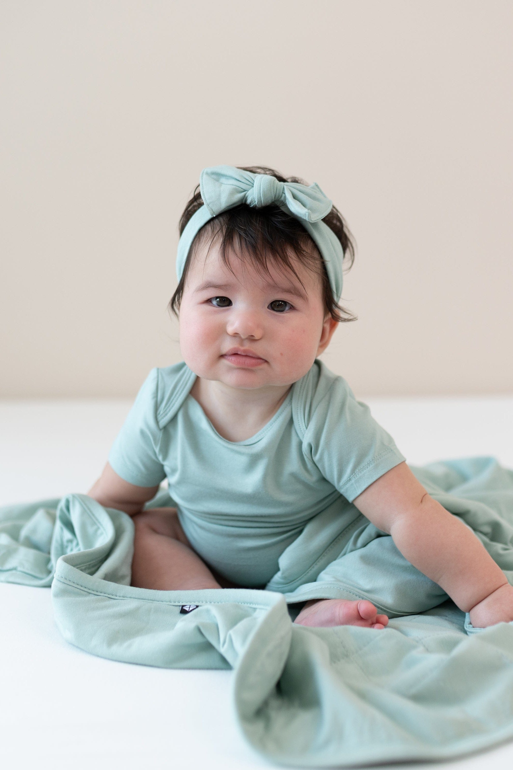Baby sitting on a Baby Blanket in Sage 1.0 while wearing a matching short sleeve bodysuit and headband bow