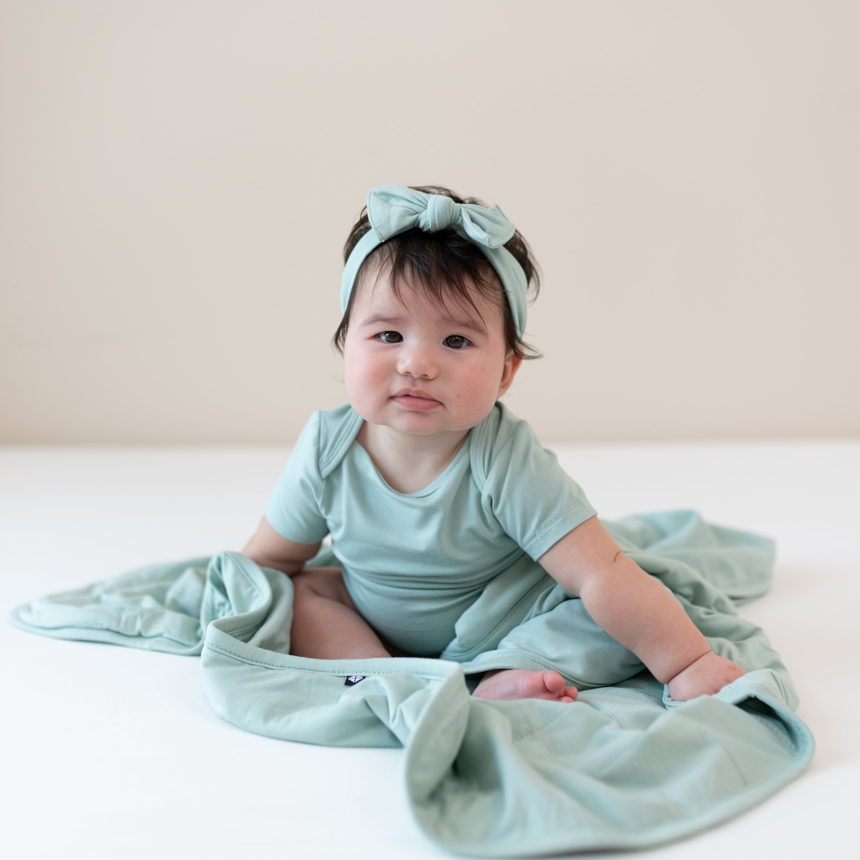 Baby sitting on a Baby Blanket in Sage 1.0 while wearing a matching short sleeve bodysuit and headband bow