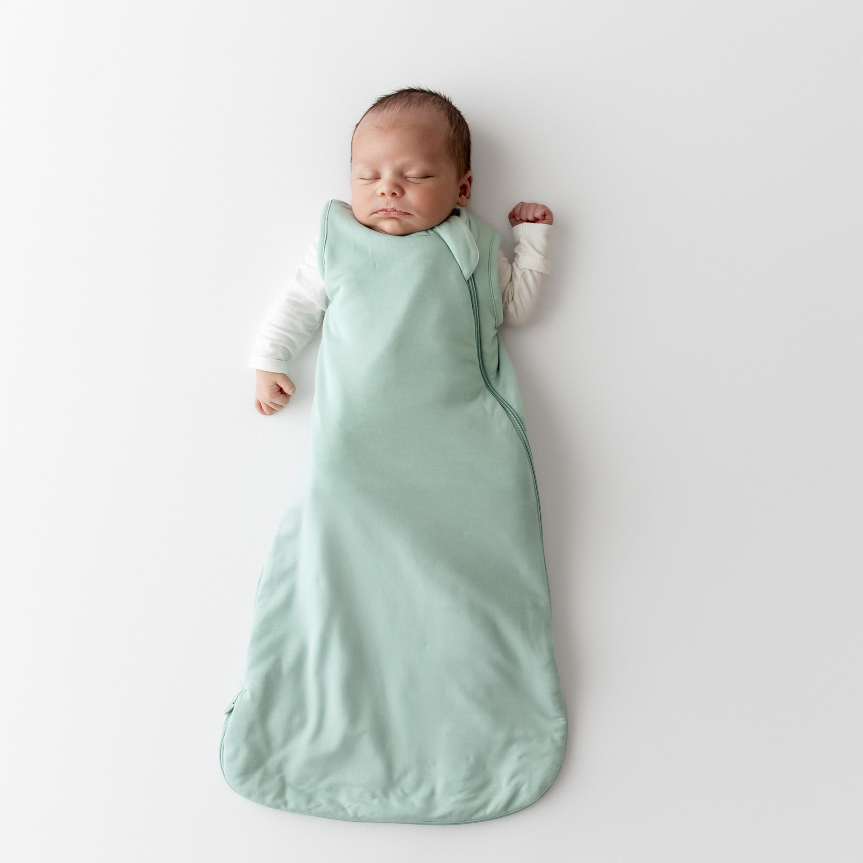 Newborn baby sleeping in a light green kyte baby sleep bag with white onesie underneath on a white background