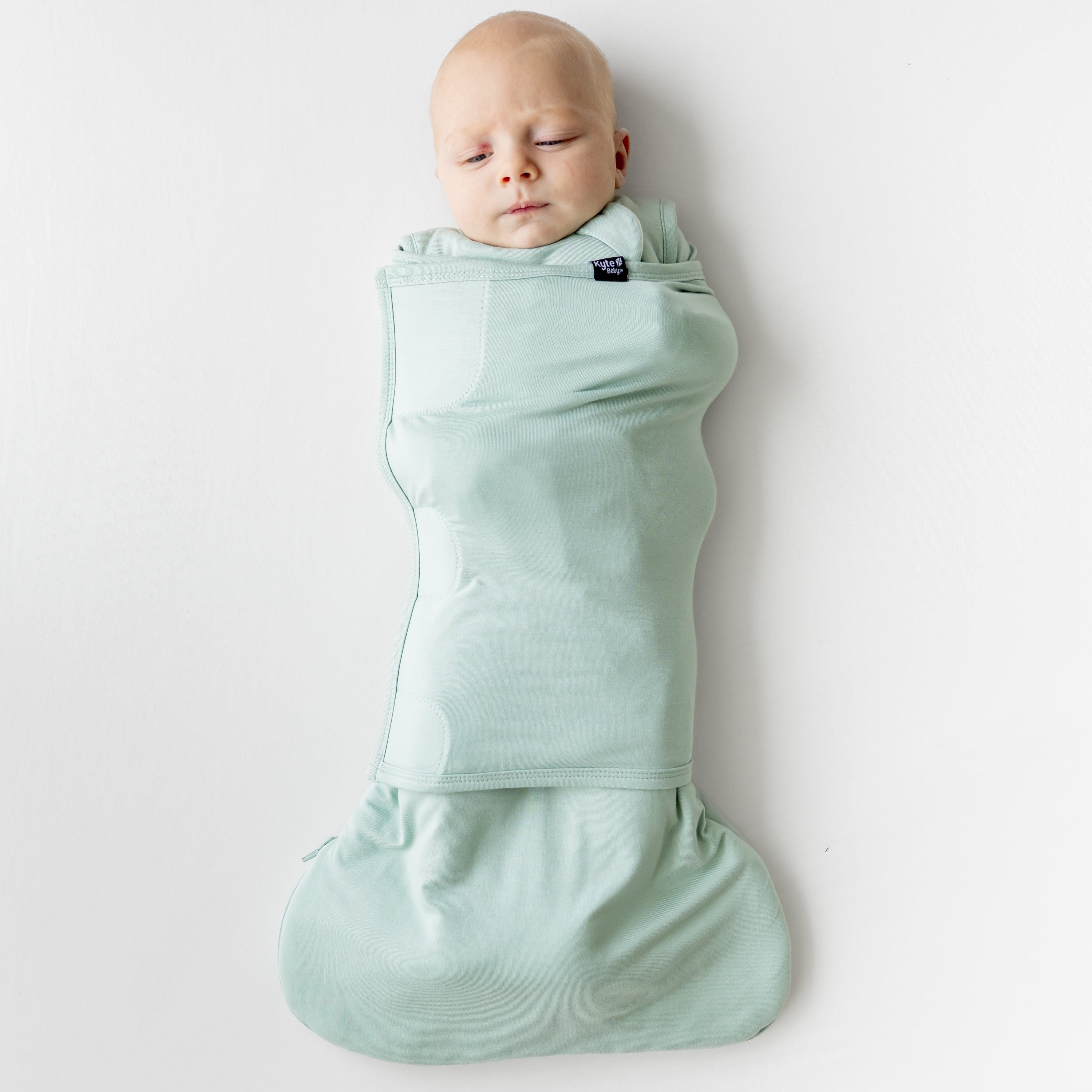 Infant laying down on a light neutral background swaddled in the Sleep Bag Swaddler in Sage 1.0
