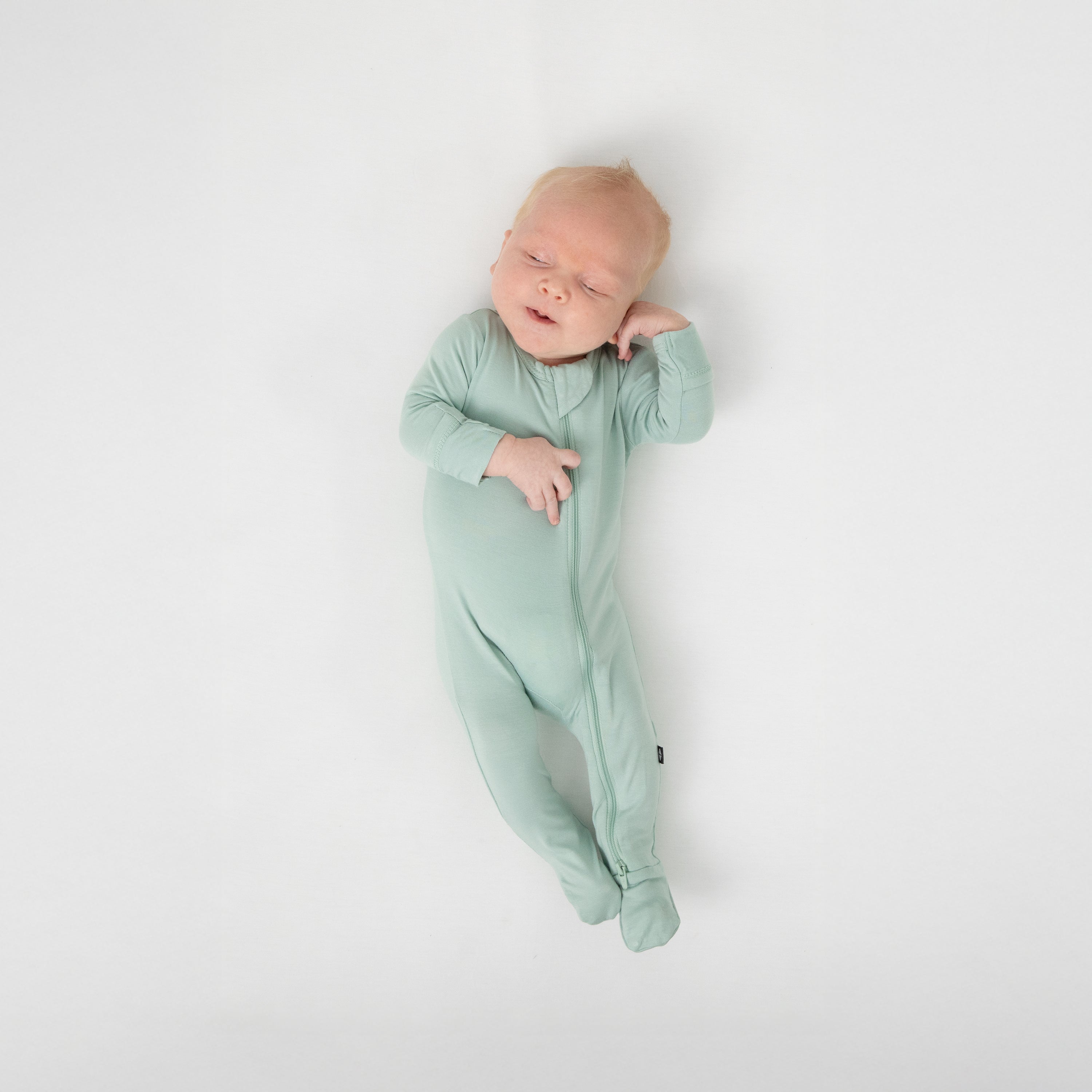 Sleeping infant laying on a white crib sheet wearing the Zippered Footie in Sage