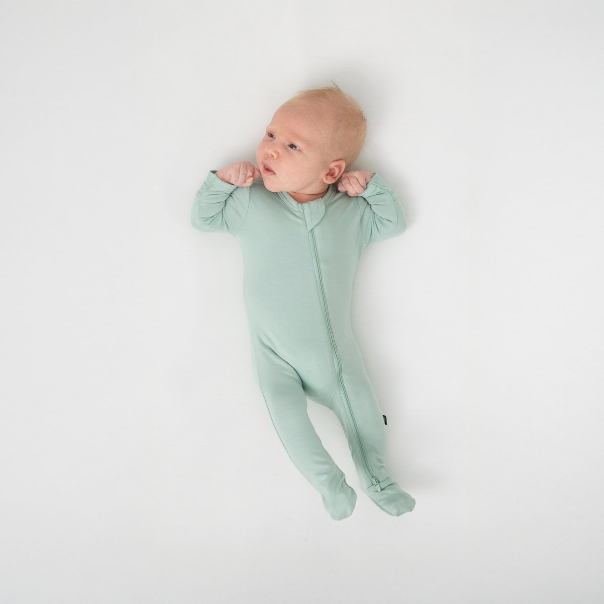 Newborn laying on a white crib sheet wearing the Zippered Footie in Sage