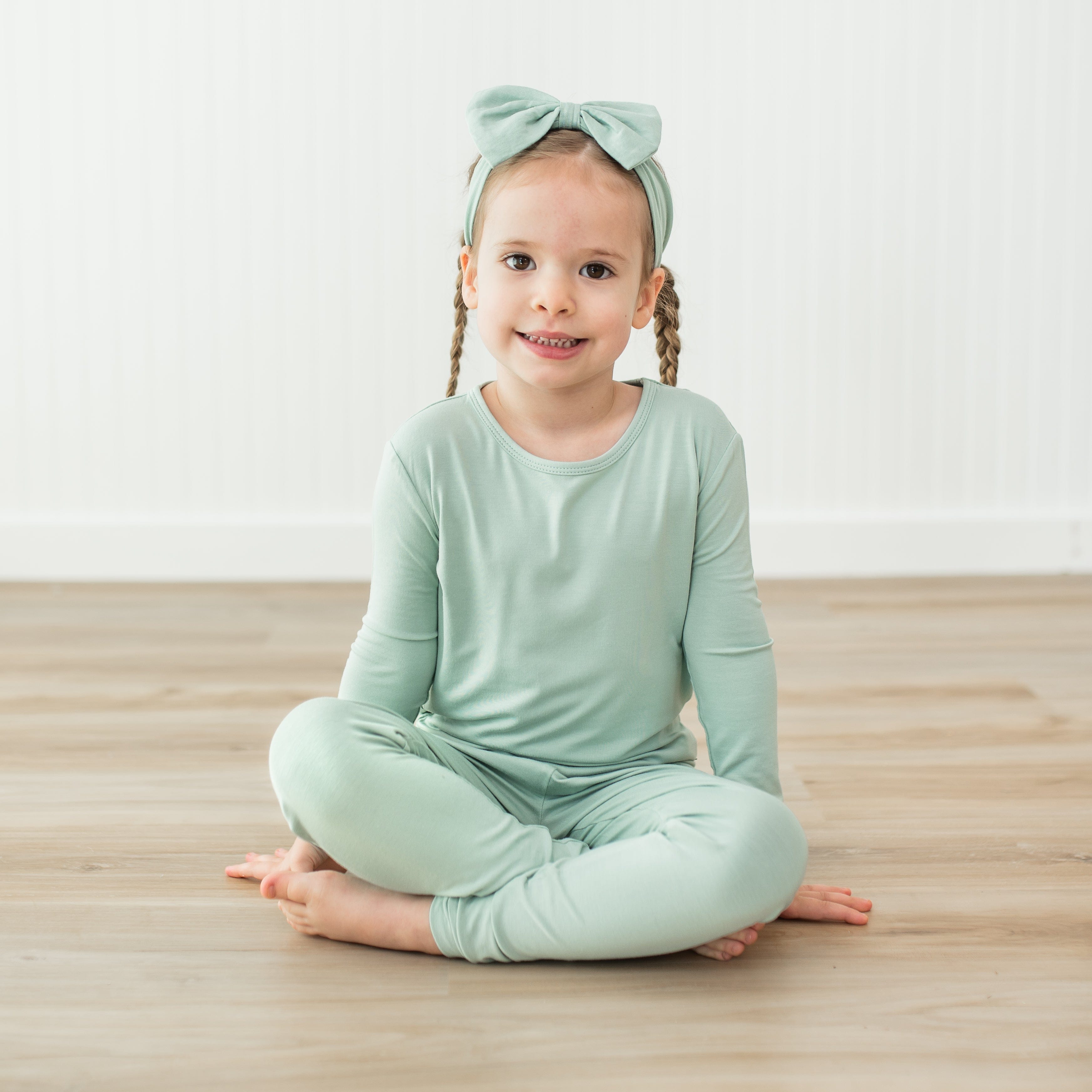 Child wearing a light green pajama with a matching headband sitting on a wooden floor.