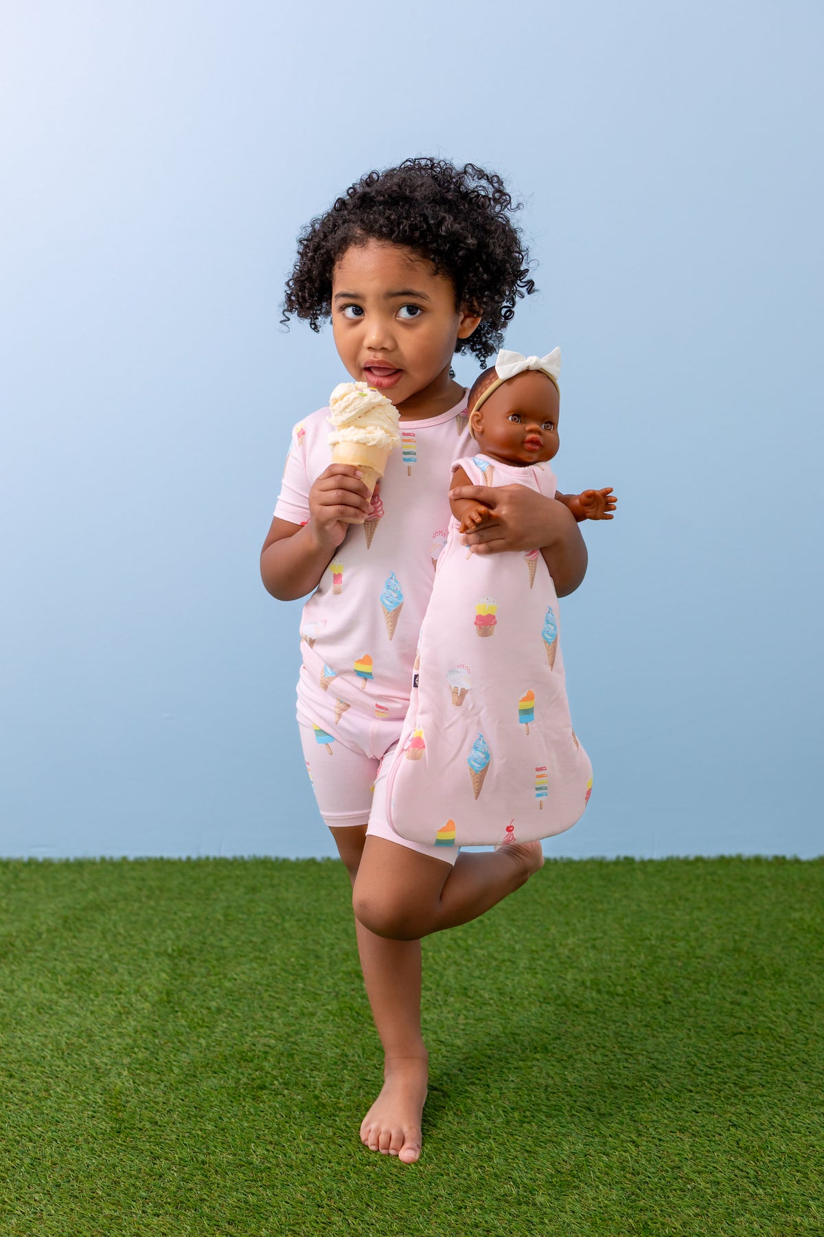 Child holding Baby doll sleep bag in Sakura Ice Cream print eating an Ice cream cone wearing a similar print Short sleeve pajama set on green grass