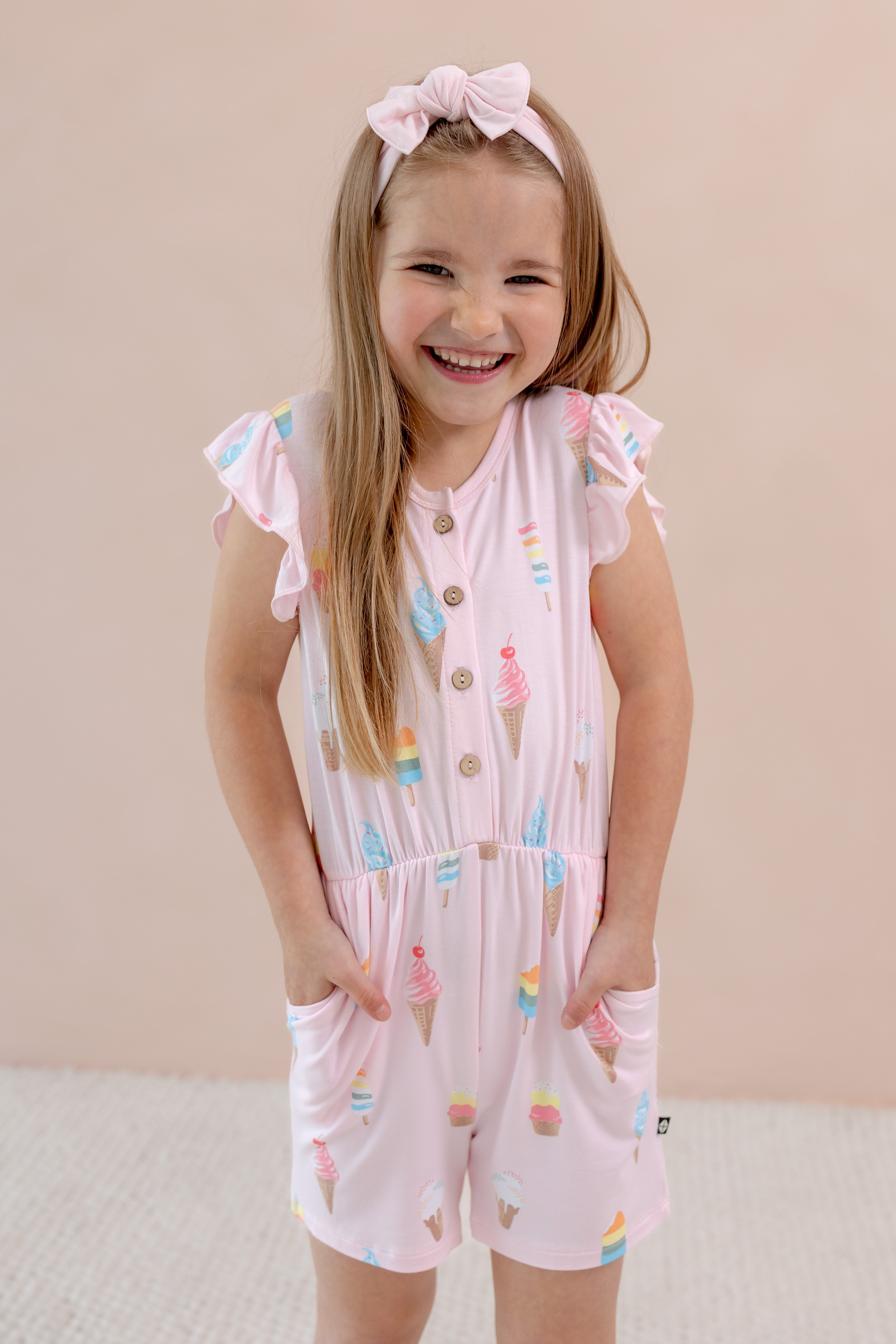 Child posing in Sakura Ice cream print Flutter Sleeve Romper with pockets on a beige background wearing a pink bow headband