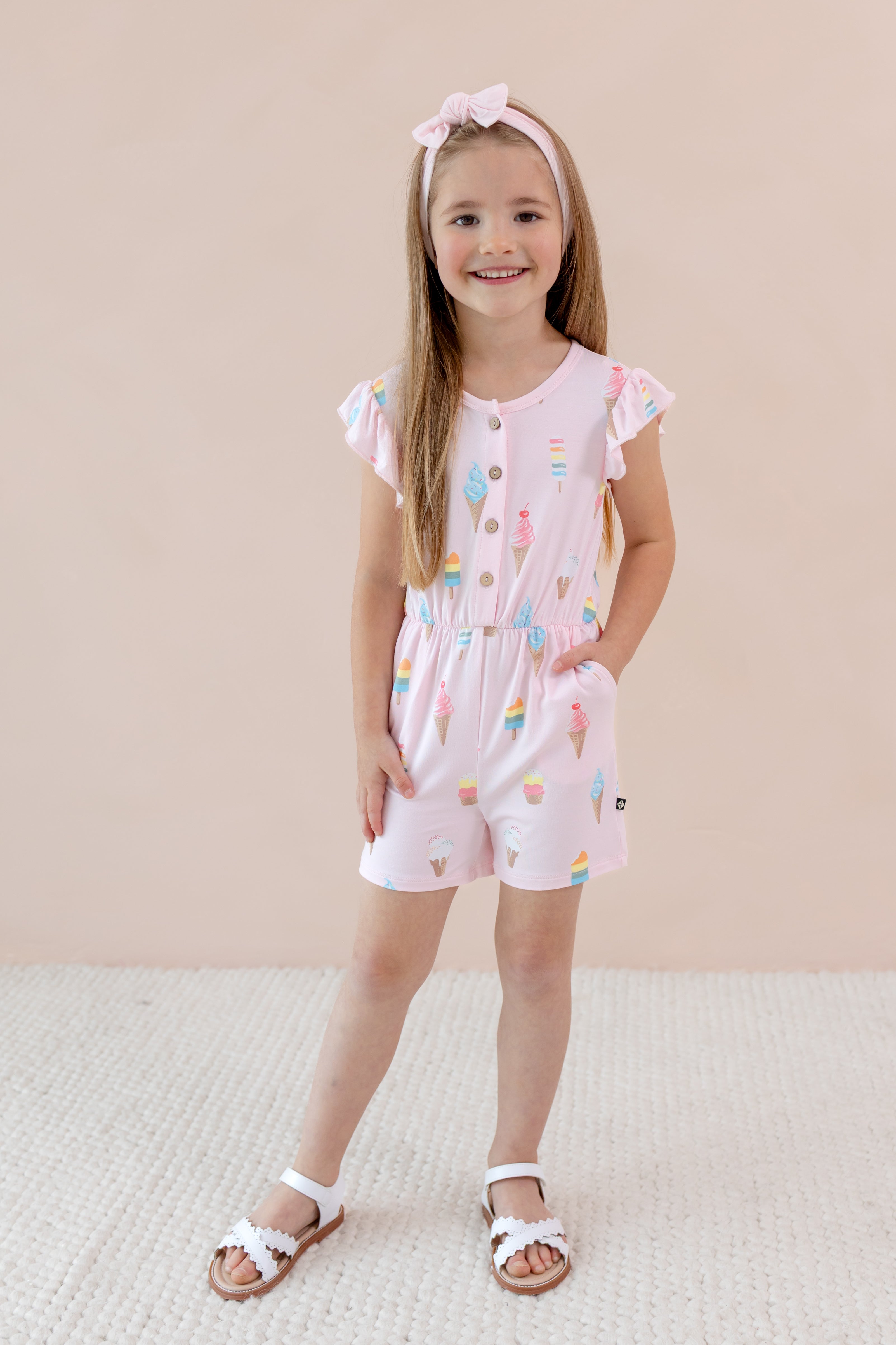 Child standing in Flutter Sleeve Romper in Sakura Ice Cream print with pink bow head band and white sandles on a beige background