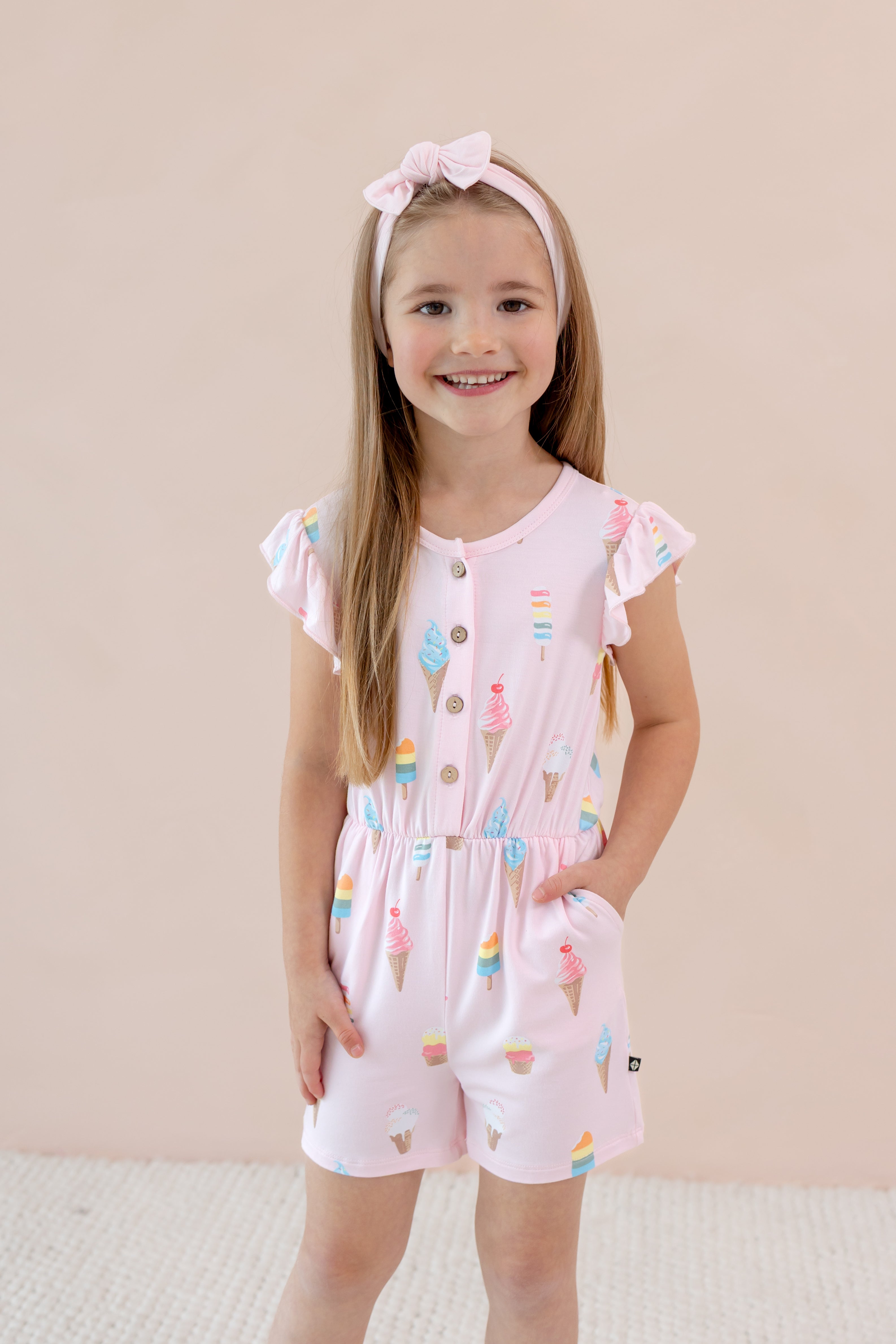 Child wearing a Flutter Sleeve Romper in Sakura Ice Cream print with pink bow head band on a beige background