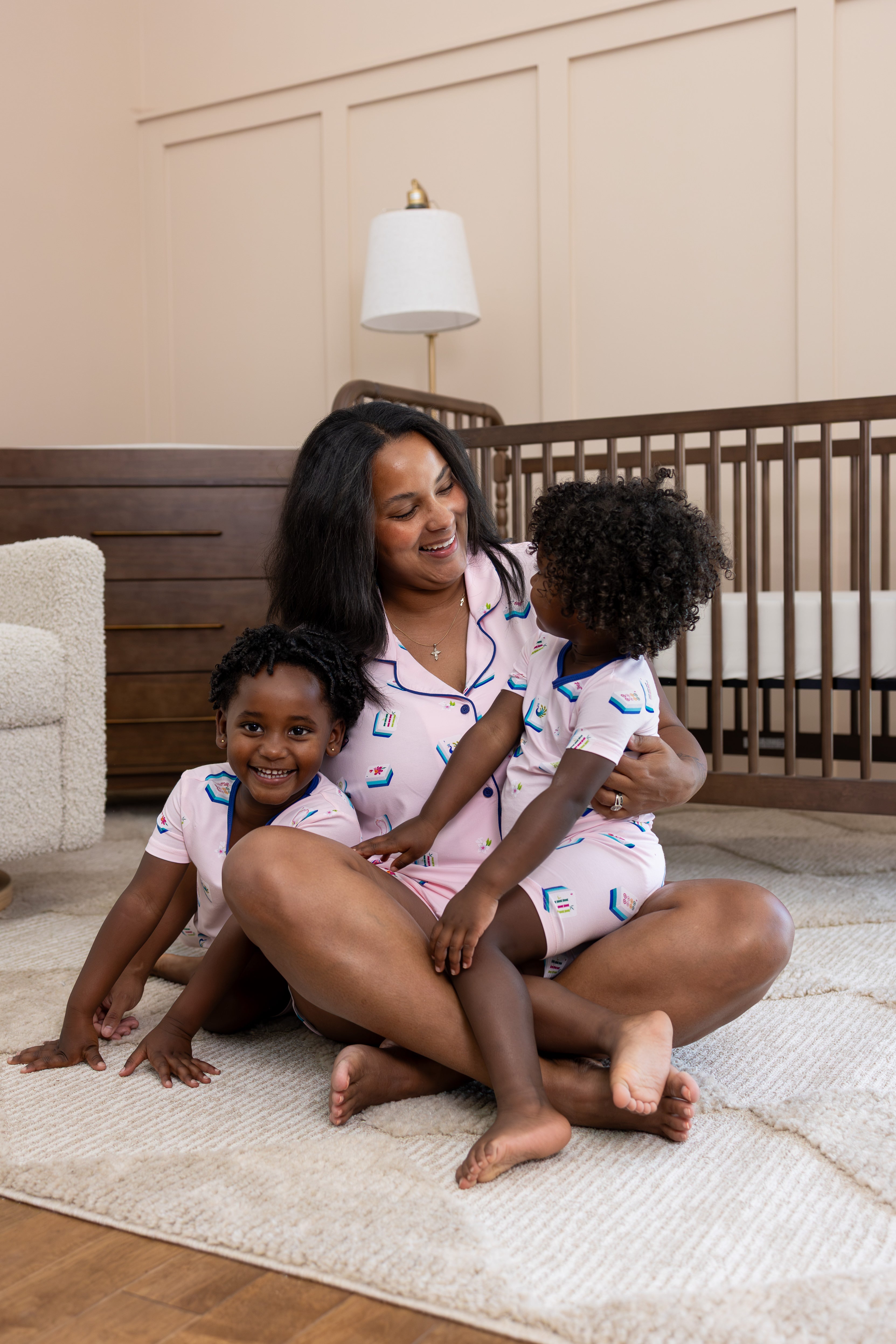 A woman and two young children wearing matching light pink pajamas with a blue mahjong pattern are sitting together on a cream rug in a nursery setting.