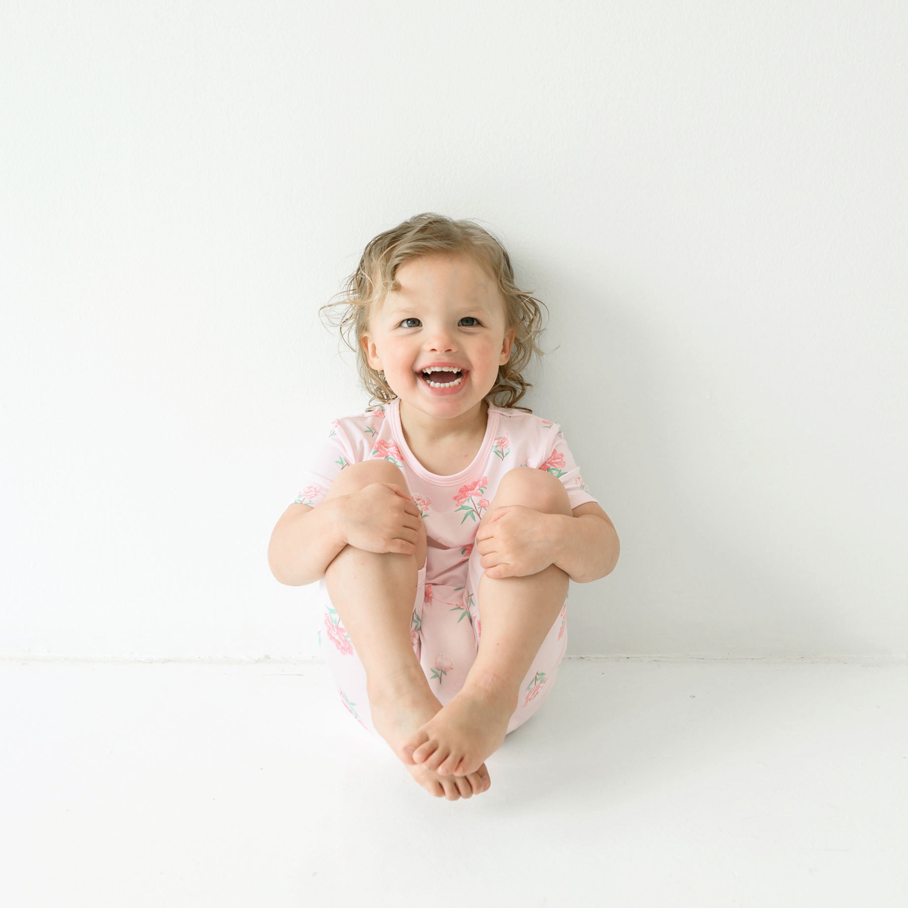 Smiling toddler model sitting with her legs up wearing the Short Sleeve Pajamas in Sakura Peony