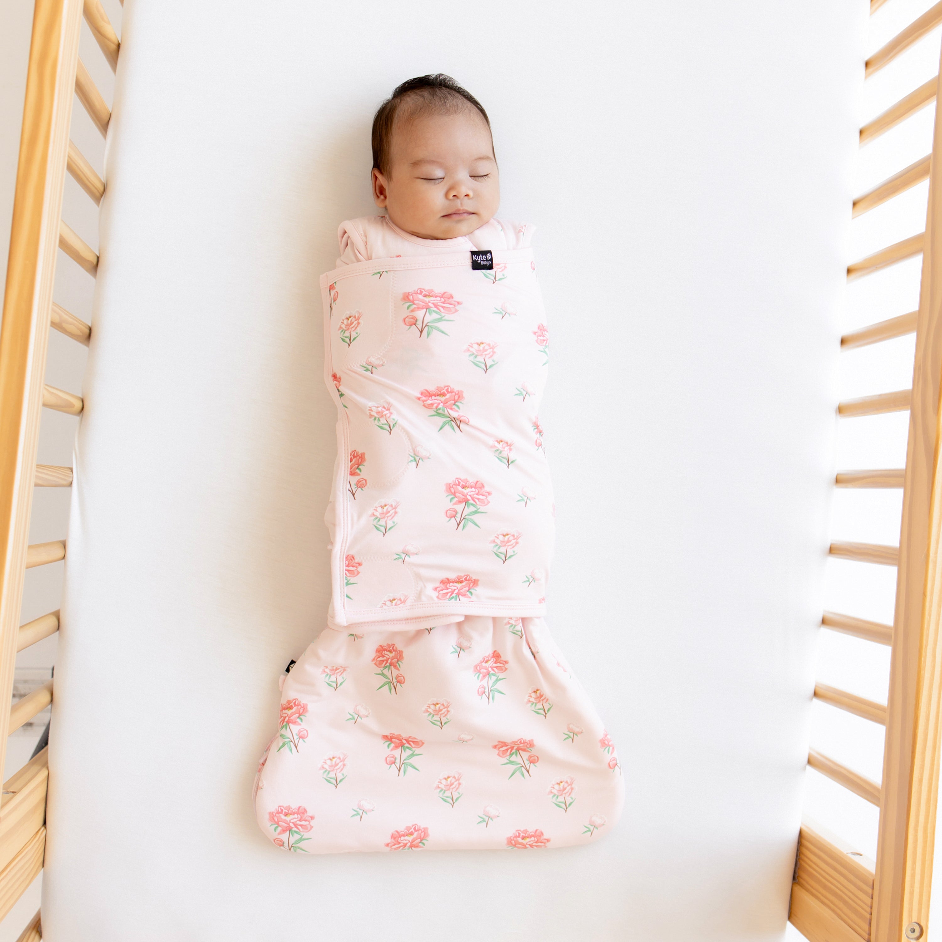 Sleeping baby shown swaddled in the Sleep Bag Swaddler in Sakura Peony 1.0 laying in a crib on a Cloud Crib Sheet