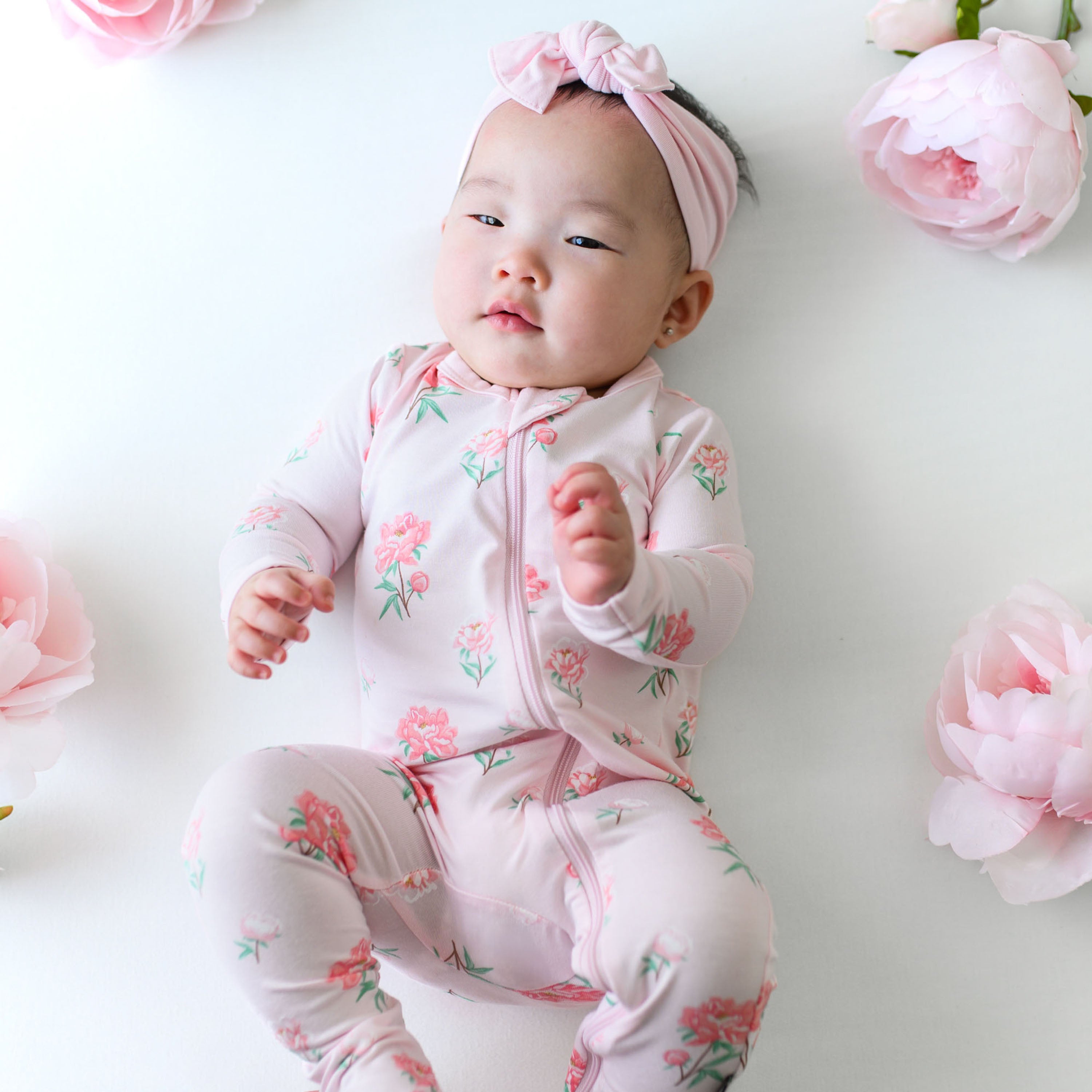 Infant wearing pink Zippered Footie pajamas in Sakura Peony and Knotted Bow headband