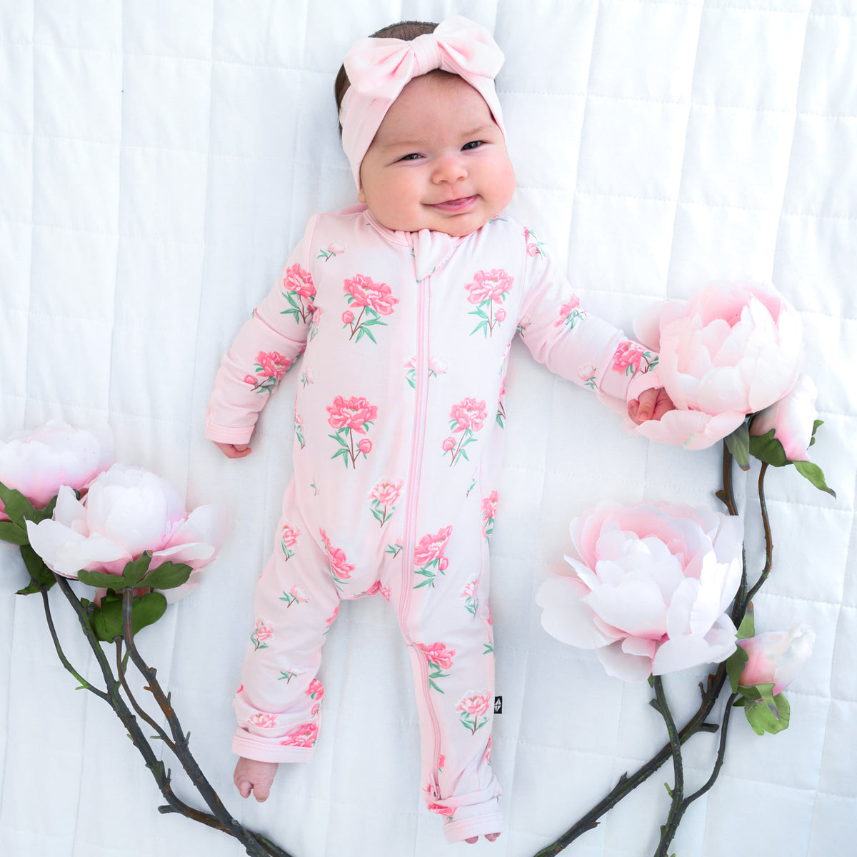 Smiling newborn laying on a Cloud Blanket wearing the Zippered Romper in Sakura Peony with artificial Peony flowers on stems around her