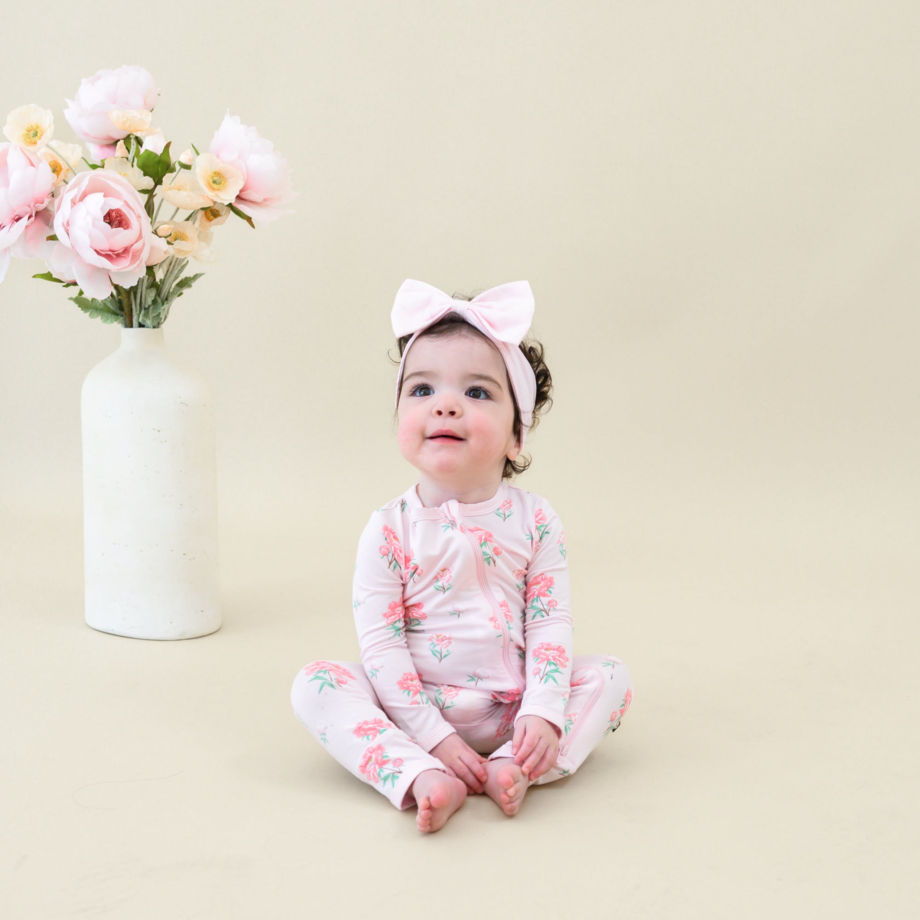 Toddler sitting beside a vase of Peony's modeling the Zippered Romper in Sakura Peony paired with a Sakura Bow Headband