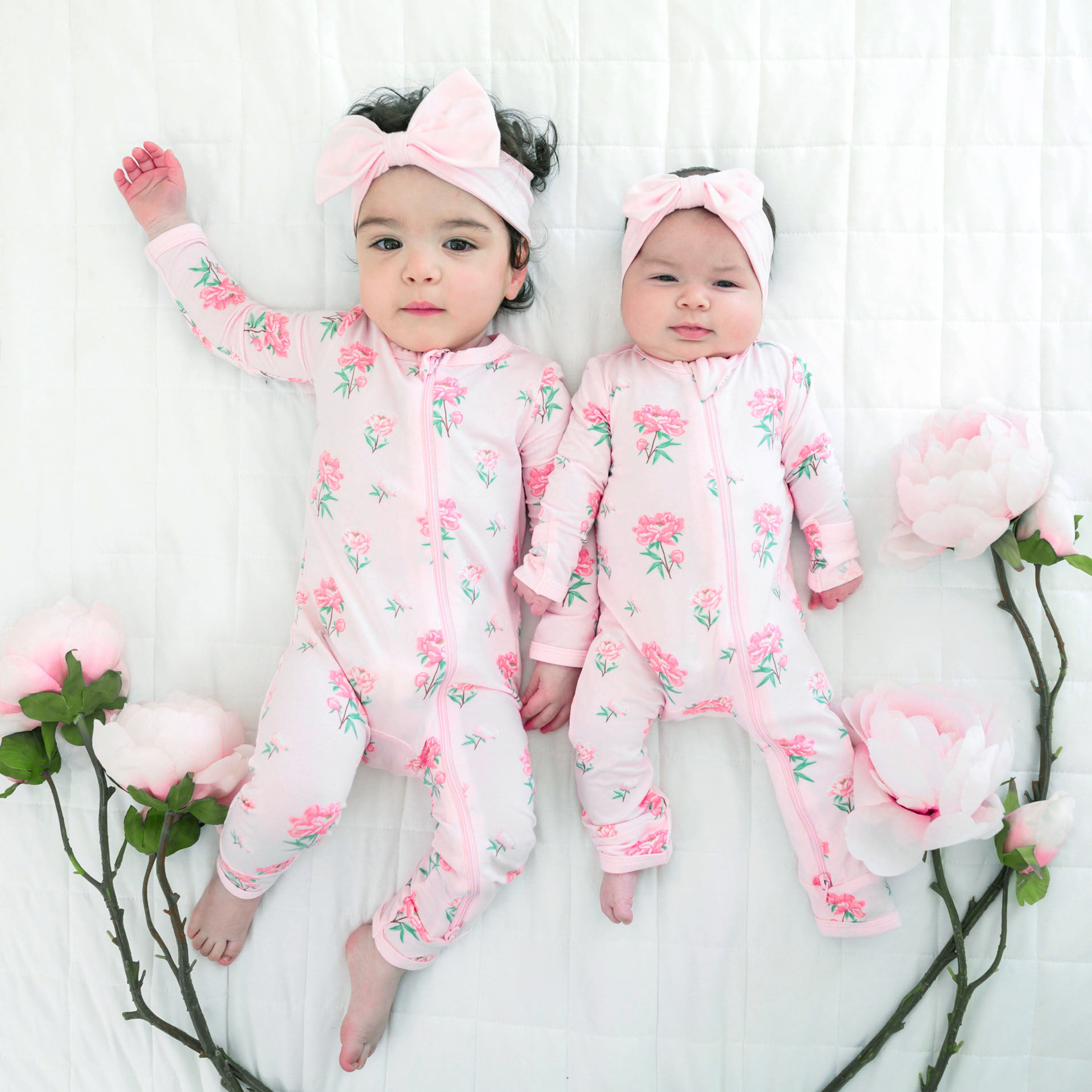 A toddler and a newborn laying side by side wearing Zippered Rompers in Sakura Peony and Bow headbands in Sakura with Peony flowers on either side of them