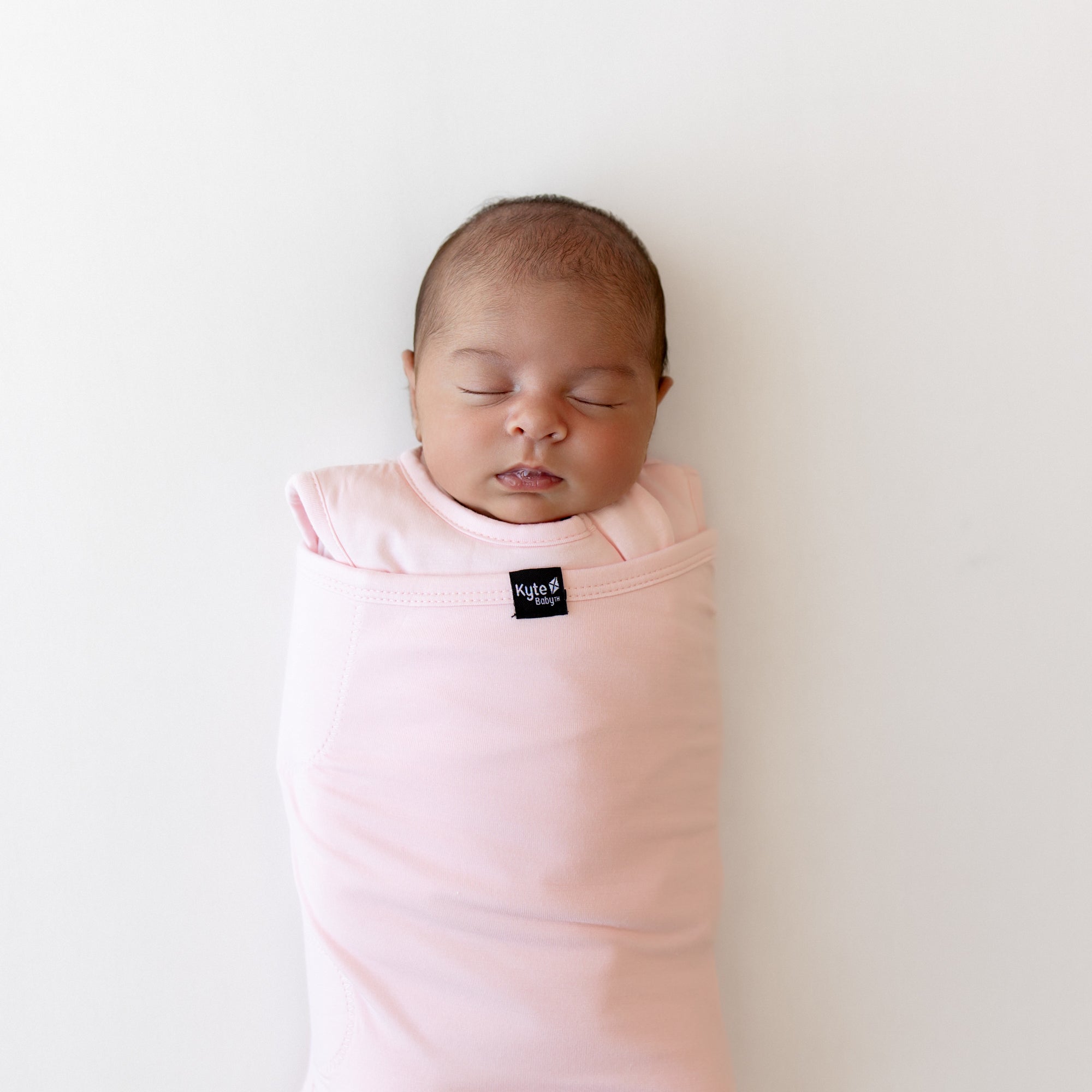Baby sleeping on a white surface wearing a sleep bag swaddler in a light pink with a kyte baby logo in center