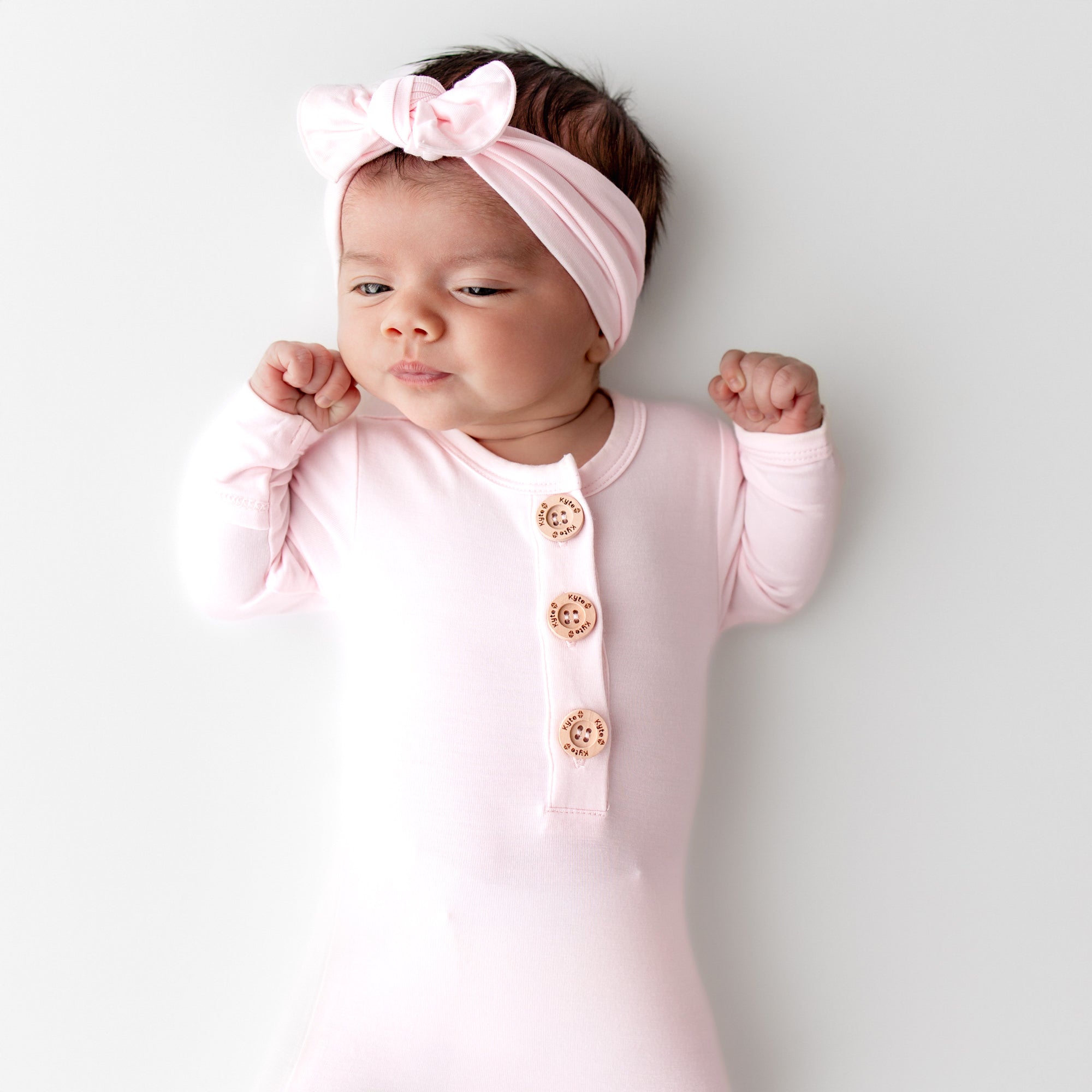 Newborn baby in a light pink knotted gown with wooden buttons and matching bow headband