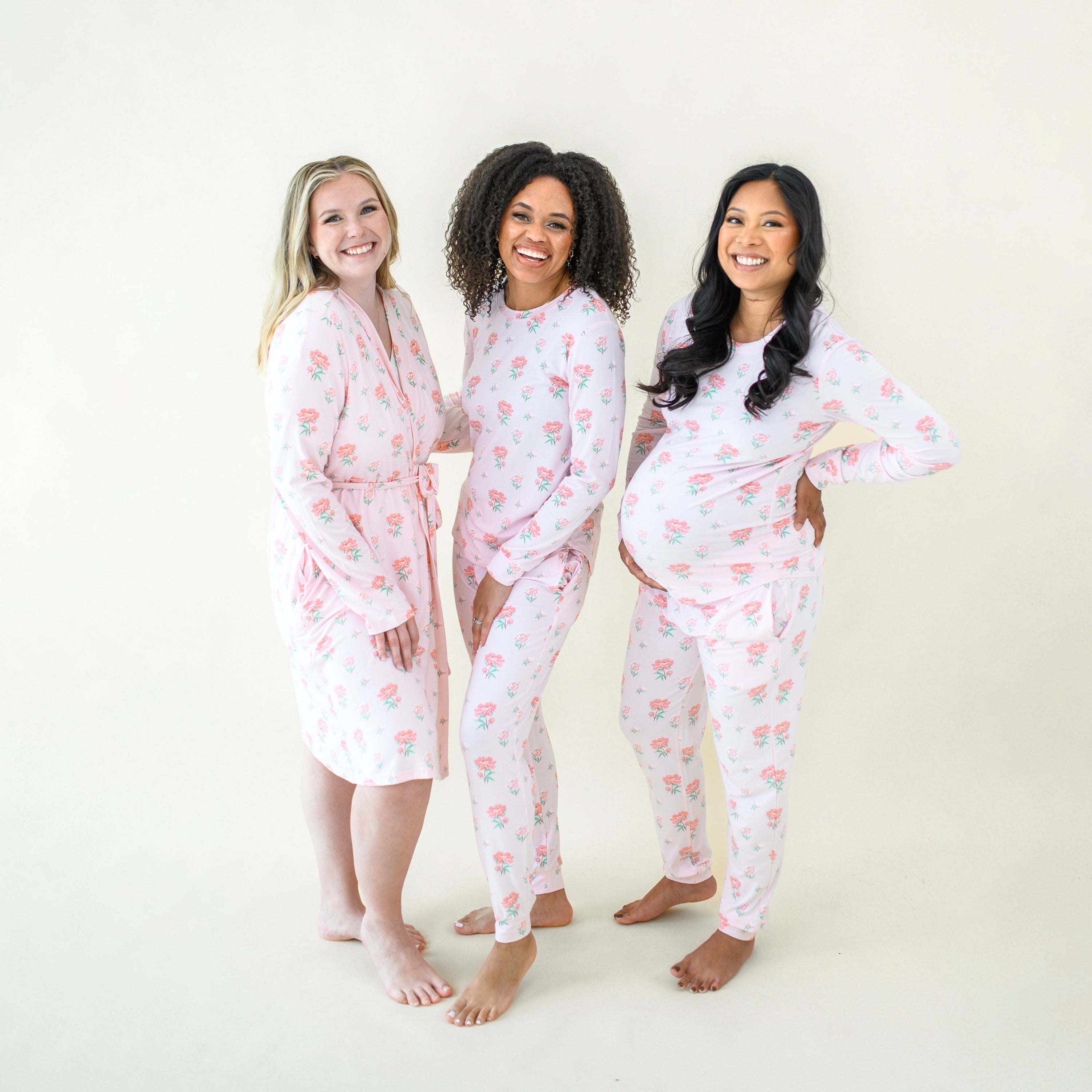 Three smiling female models wearing items from the Sakura Peony collection. The first model is wearing the women's lounge robe in Sakura Peony while the other two models are wearing the Women's Jogger Pajama Set in Sakura Peony
