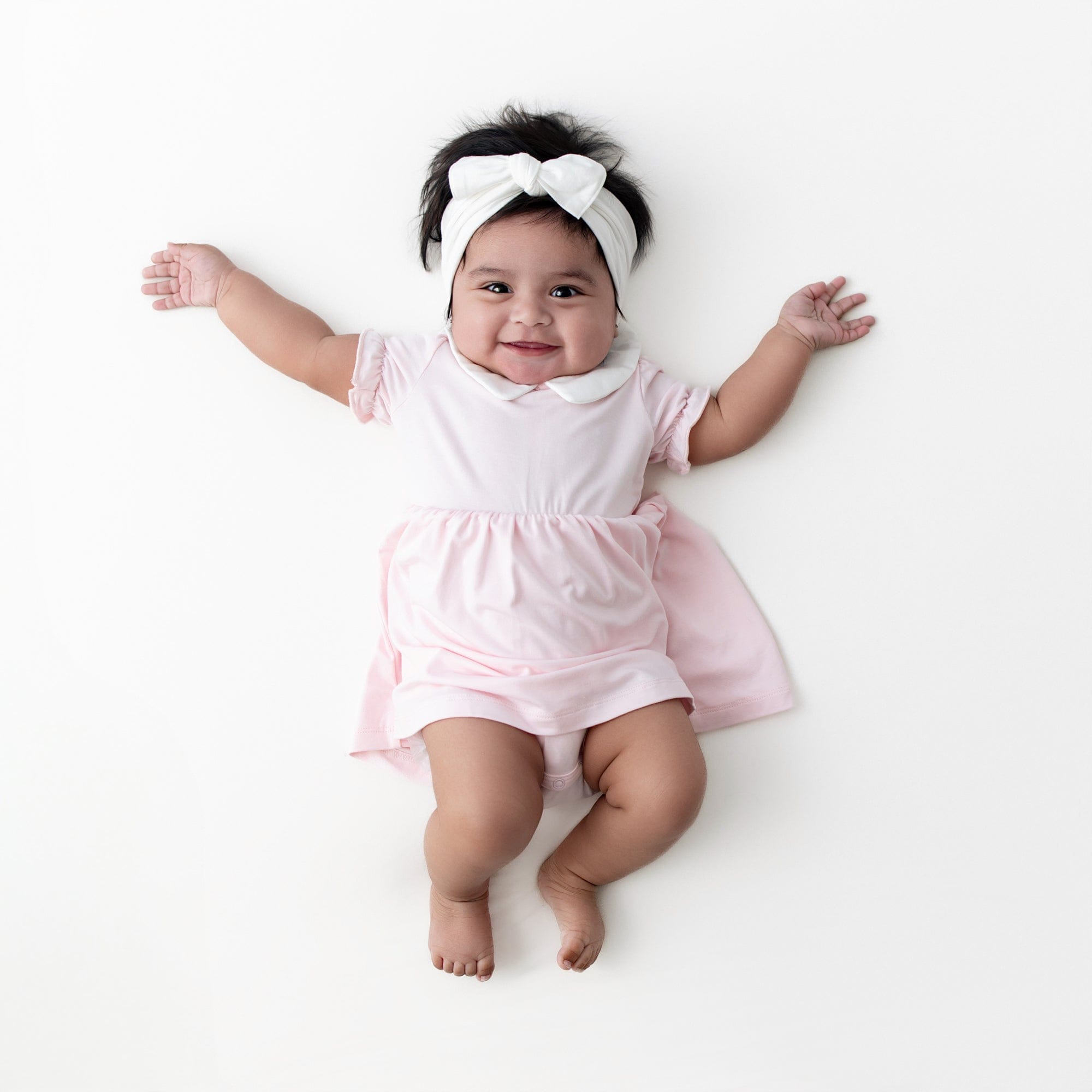 Infant girl modeling the Peter Pan Collar Bodysuit Dress in Sakura paired with a white knotted bow