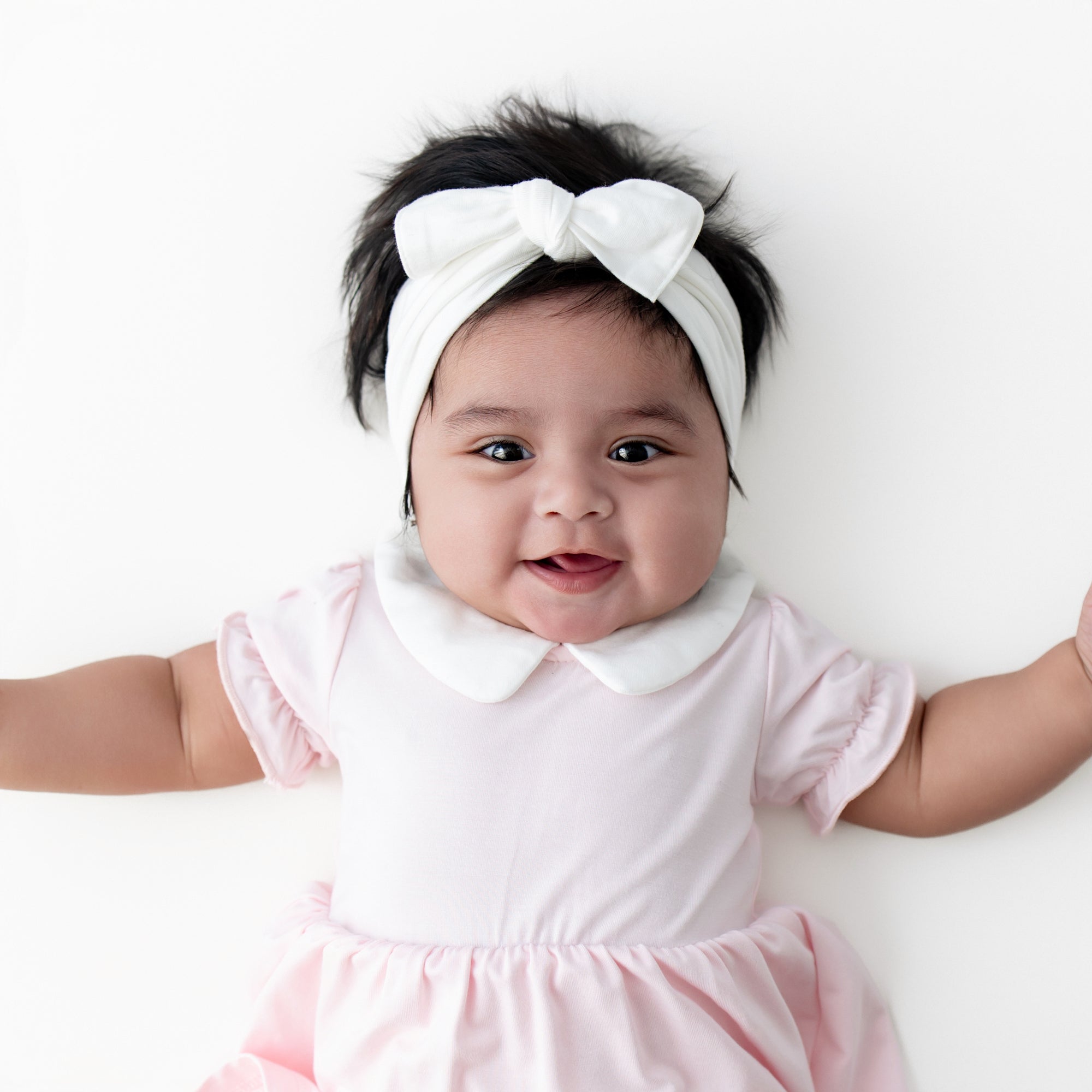 Close up of an infant girl wearing the Peter Pan Collar Bodysuit Dress in Sakura with a white knotted bow