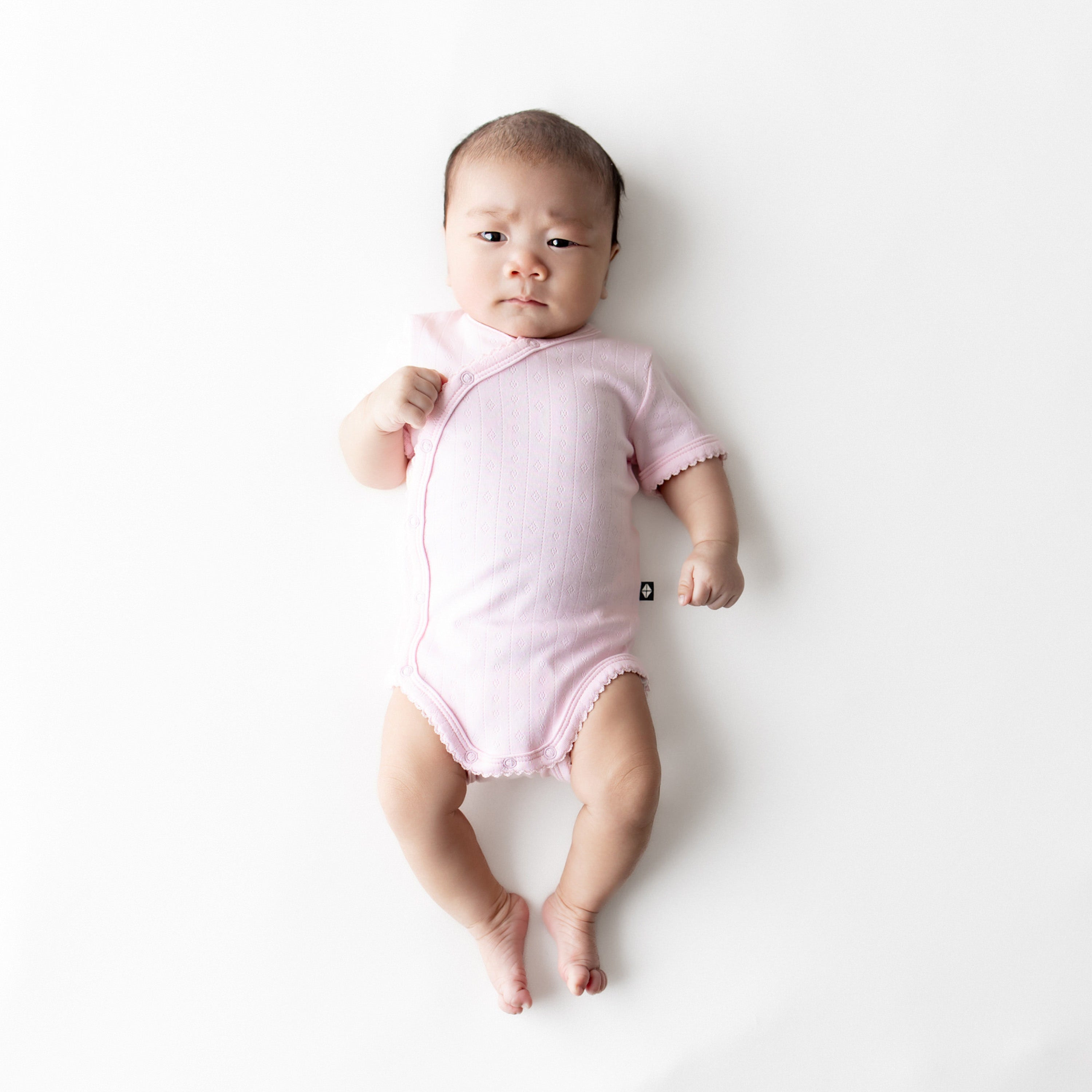 Newborn laying on a white surface wearing the Pointelle Kimono Bodysuit in Sakura with the picot trim visible