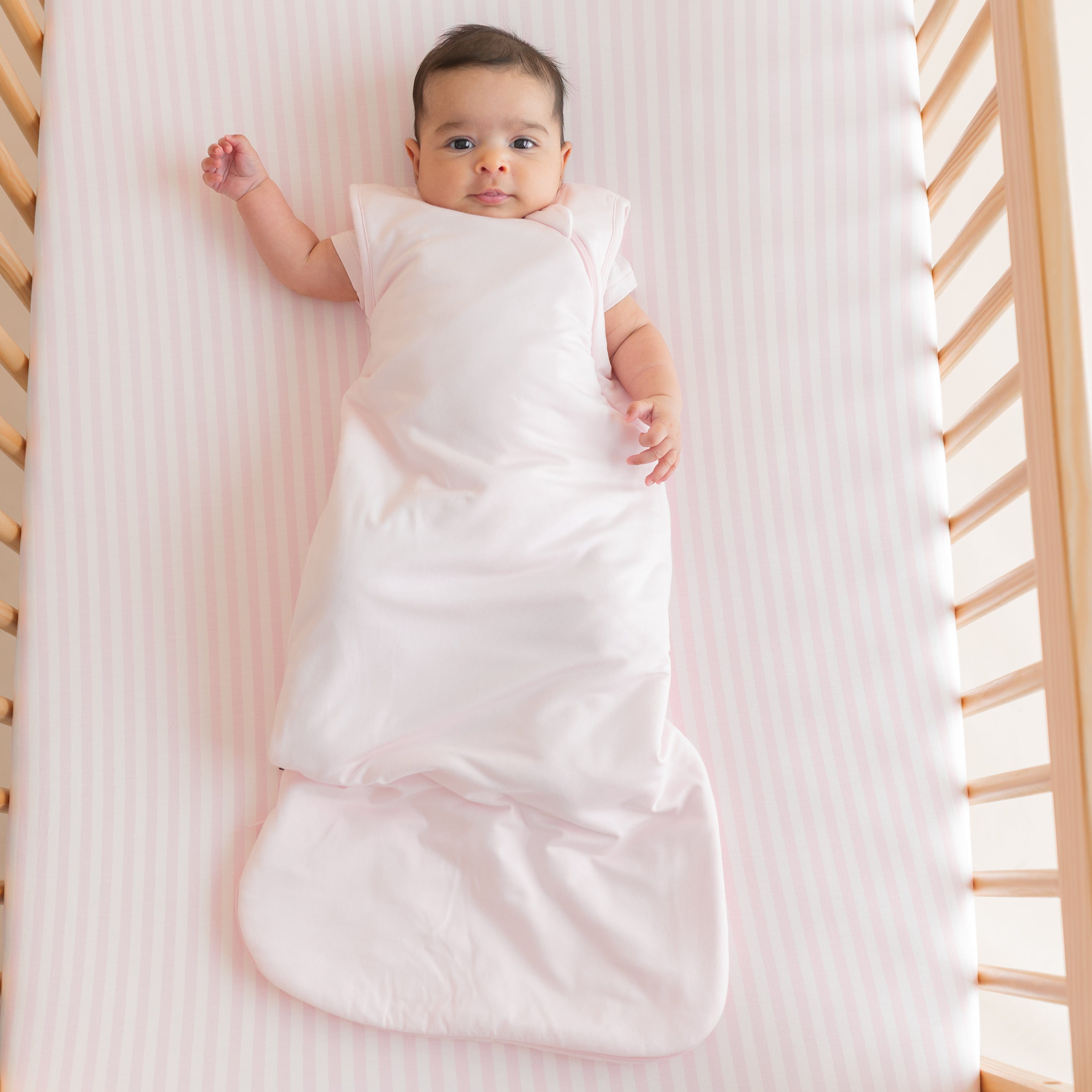 Infant laying in a crib on the Crib Sheet in Small Sakura Stripe wearing a Sakura pink sleep bag