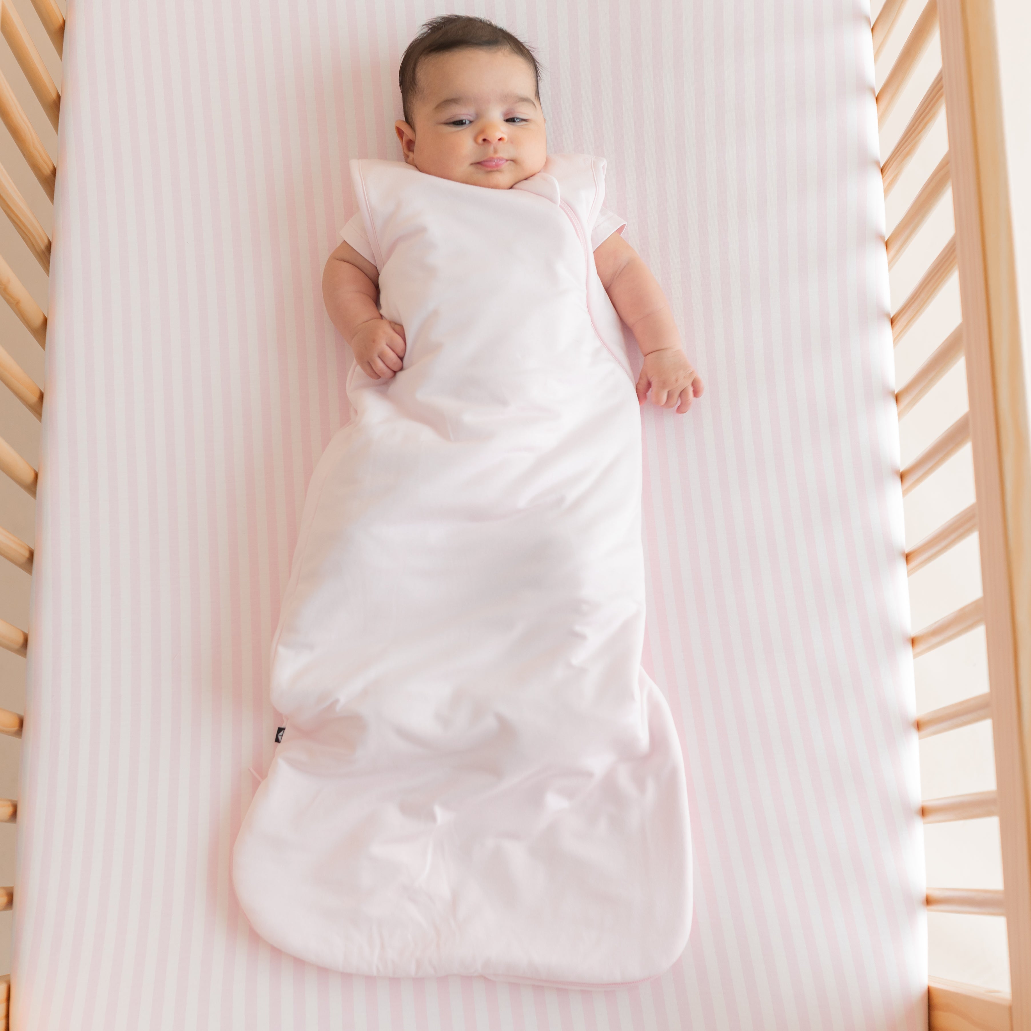 Infant laying in a crib on the Crib Sheet in Small Sakura Stripe wearing a pink sleep bag