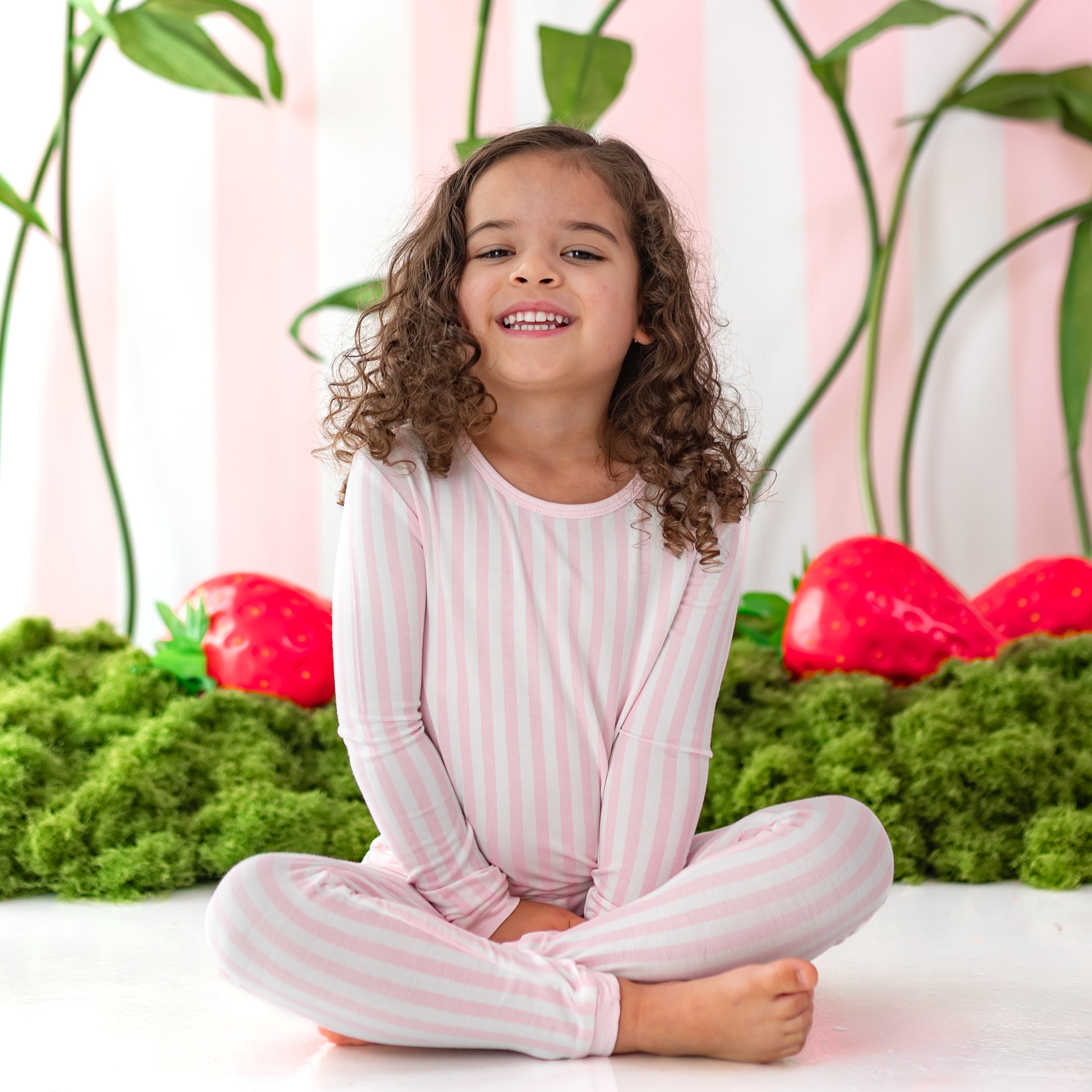 Young girl sitting on the floor in front of a strawberry backdrop wearing the Long Sleeve Pajamas in Small Sakura Stripe