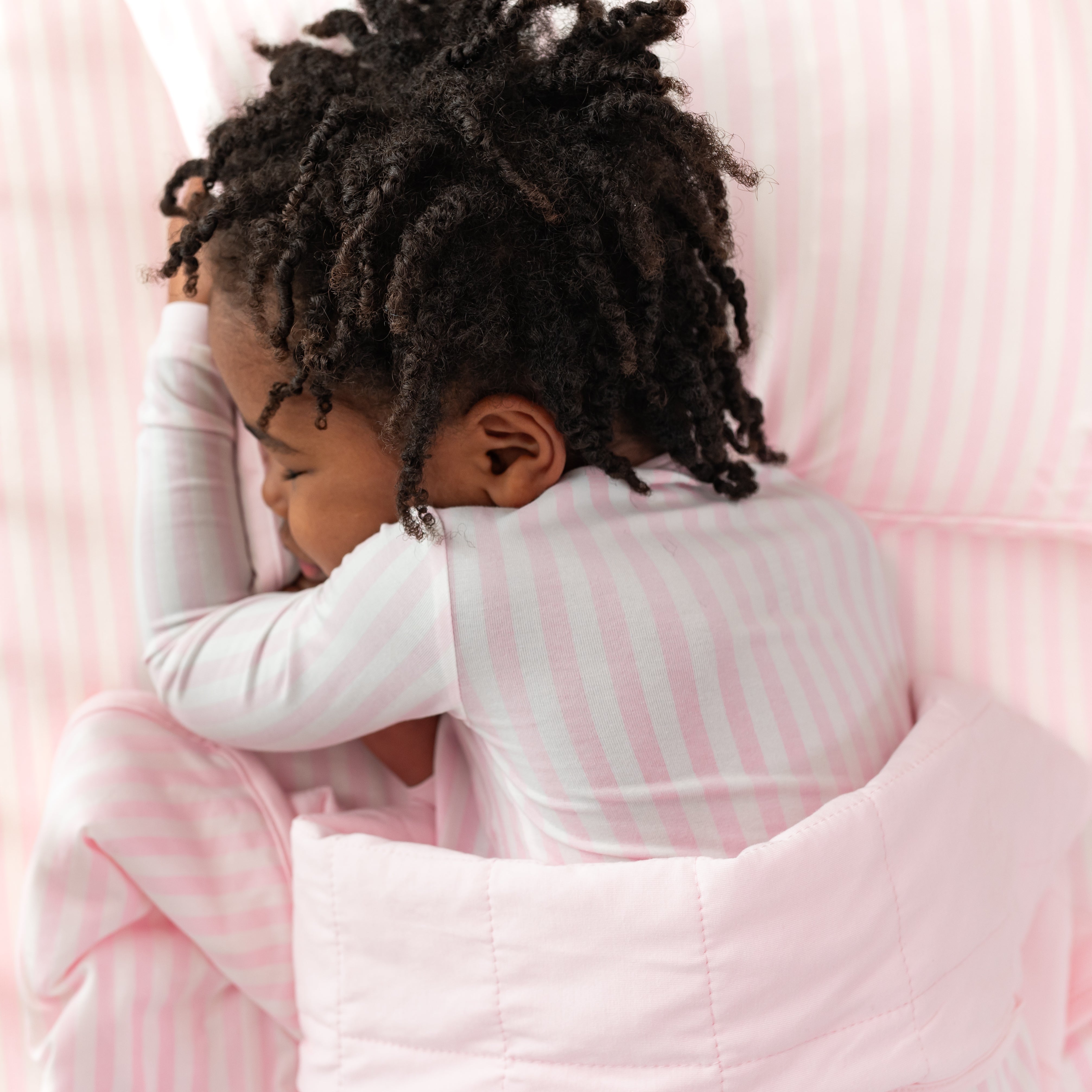 Young girl laying in bed wearing the long sleeve pajamas in Small Sakura Stripe with her head on the Toddler Pillowcase in Small Sakura Stripe underneath the matching toddler blanket