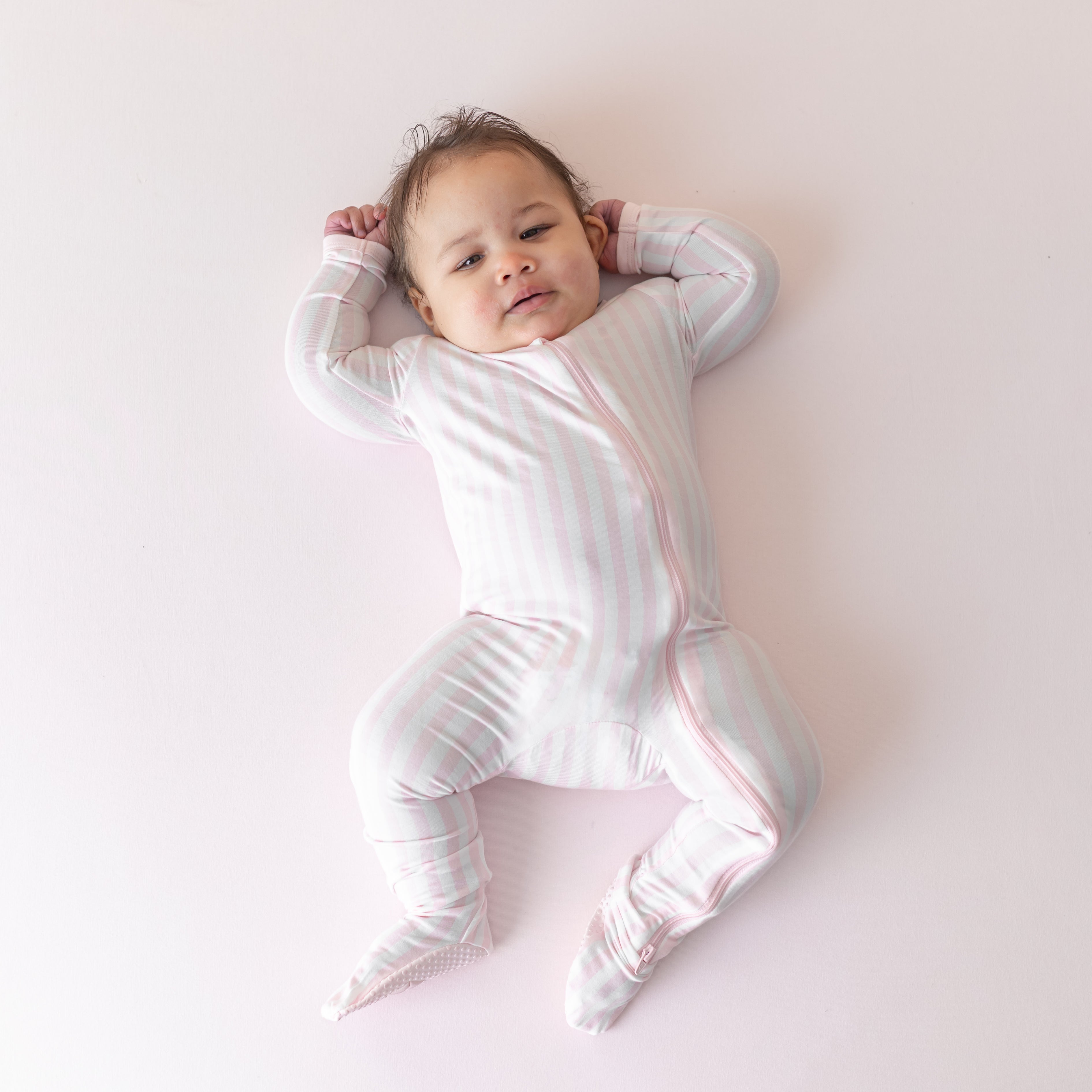 Infant laying on a light pink surface wearing the soft and breathable Zippered Footie in Small Sakura Stripe