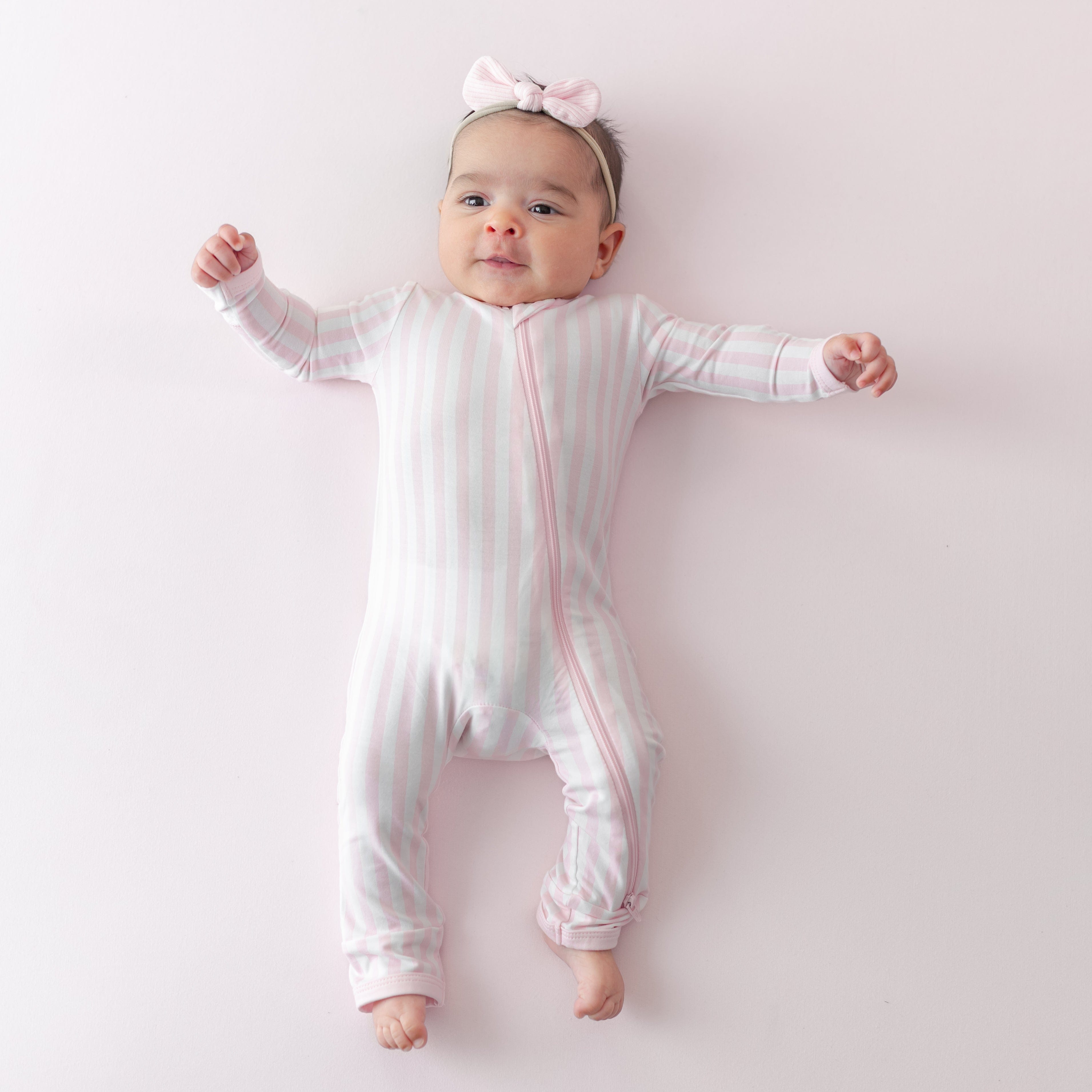 Infant laying on a light pink surface wearing the soft and stretchy Zippered Romper in Small Sakura Stripe with a pink ribbed nylon bow