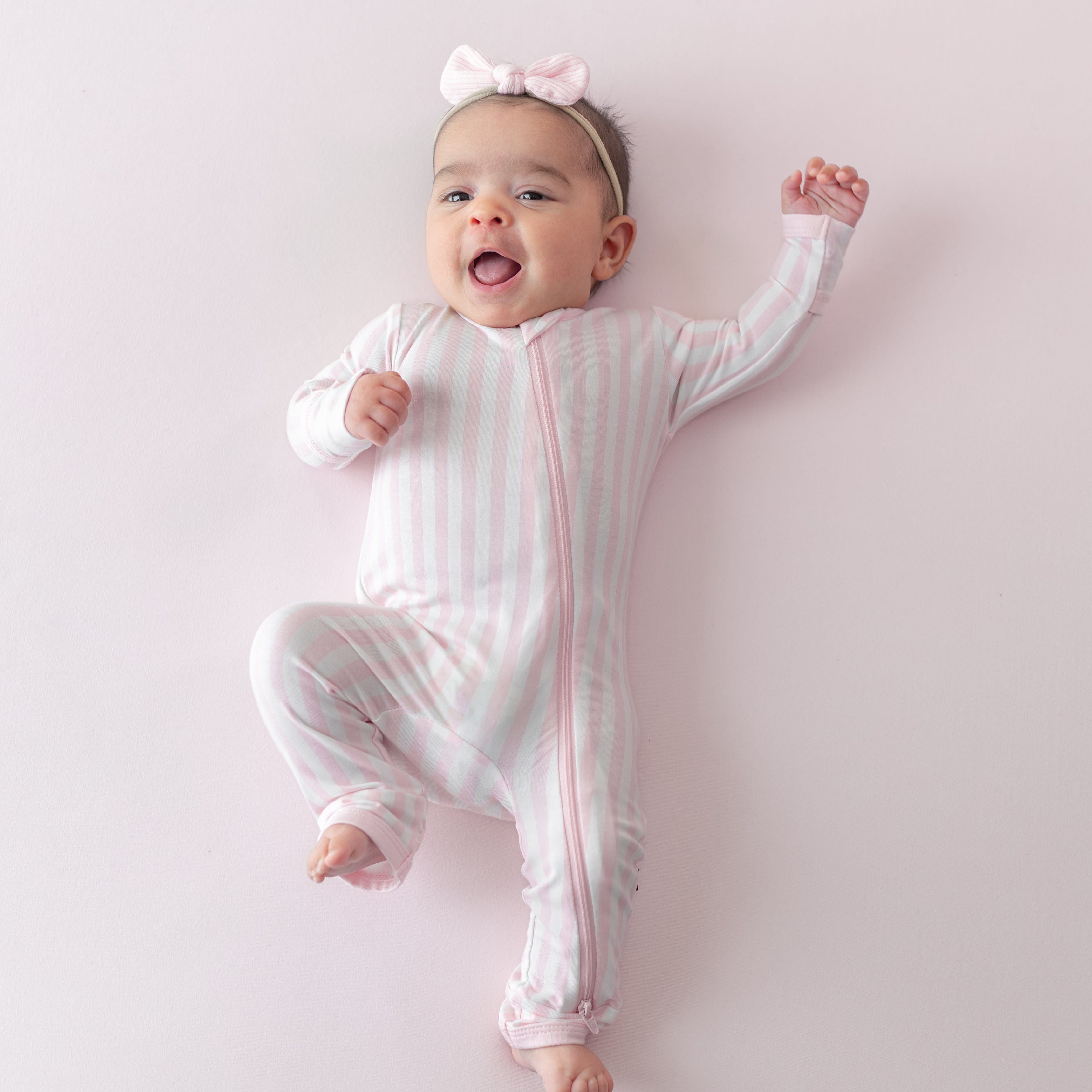 Smiling infant laying on a light pink surface wearing the Zippered Romper in Small Sakura Stripe and pink ribbed headband